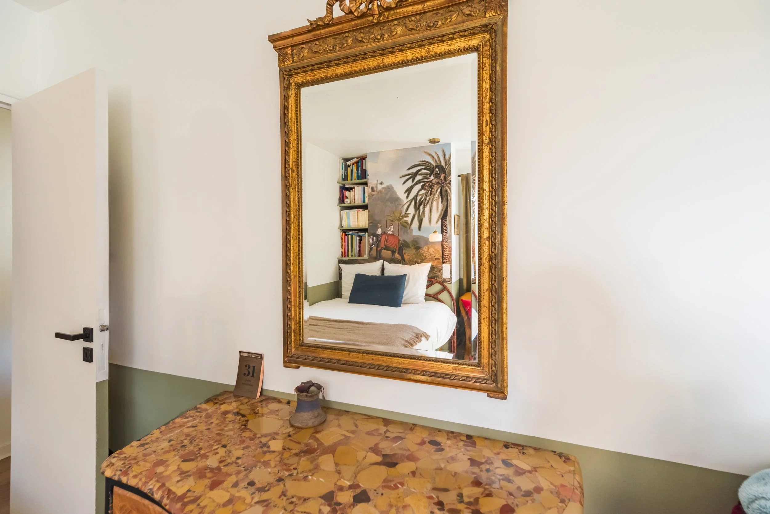 Un grand miroir encadré de bois doré accroché à un mur blanc, reflétant un coin de lit avec une tête de lit en forme de paysage avec des palmiers et une peinture murale d'une scène tropicale, une étagère avec des livres, une lampe, et une chaise colo