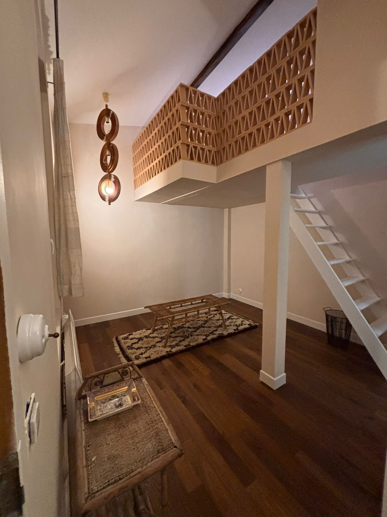 Intérieur d'une pièce avec un escalier blanc, un tapis avec un motif géométrique, un éclairage suspendu, une cheminée et un mur beige.