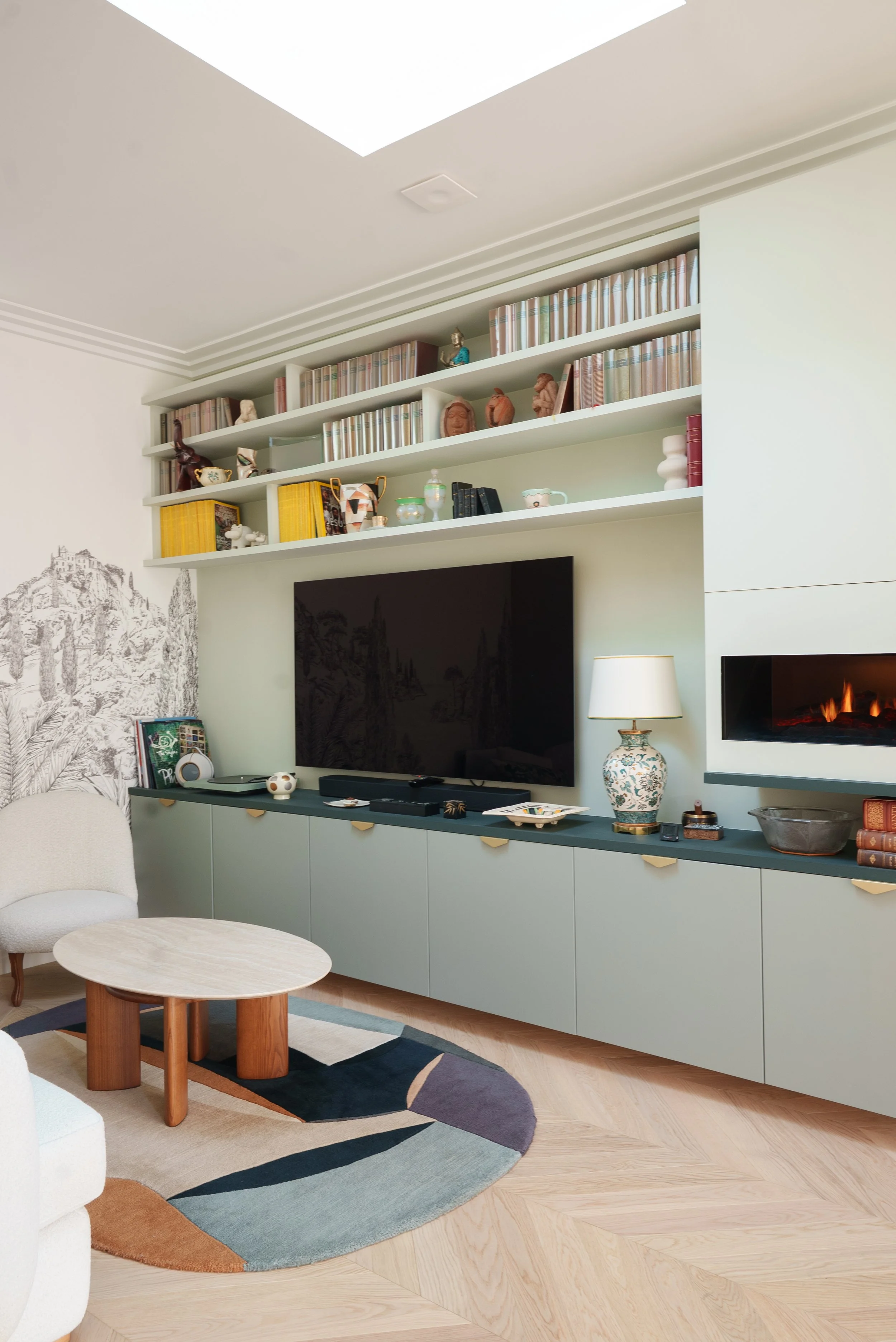 Salon avec une grande télévision, une cheminée électrique, et des étagères remplies de livres et d'objets décoratifs, comprenant une armoire de couleur vert pâle, un fauteuil blanc, une petite table centrale, un tapis coloré, et une lampe de table en