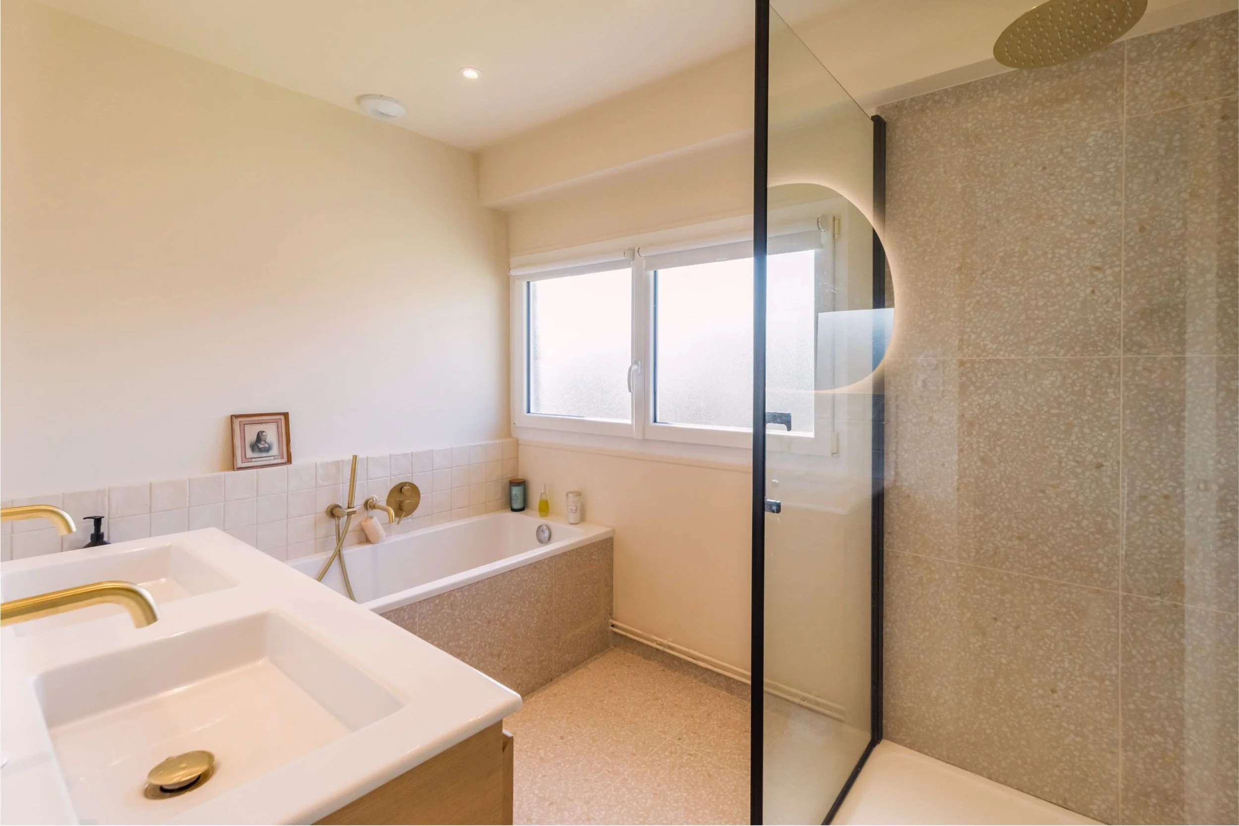 Une salle de bain avec une baignoire, un lavabo avec un comptoir en bois, un miroir rond, une douche séparée en verre avec un sol en mosaïque beige, et une grande fenêtre laissant entrer la lumière naturelle.