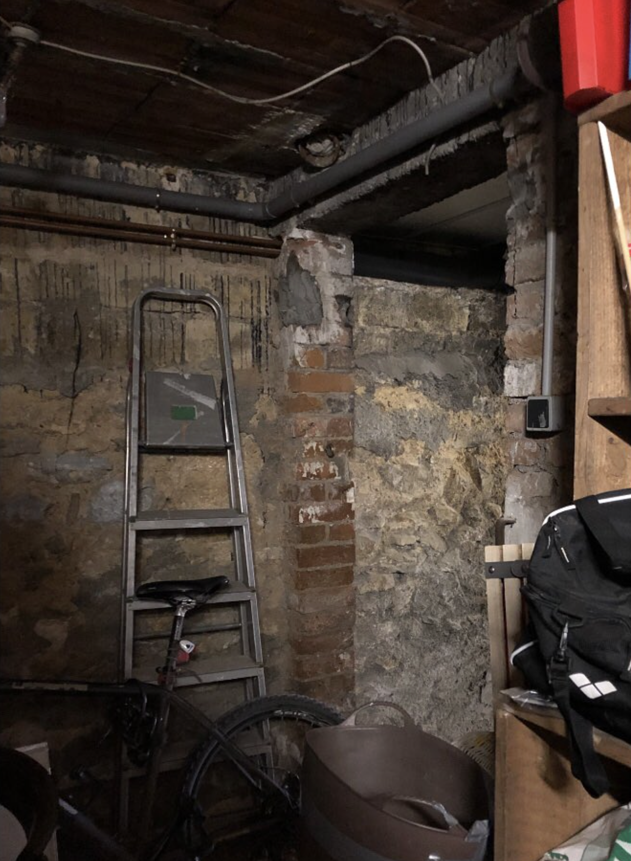 Un mur de pierre et de brique dans un sous-sol ou une cave, avec une échelle en aluminium, un vélo, une boîte en plastqiue, et un sac à dos noir placé sur une étagère en bois.