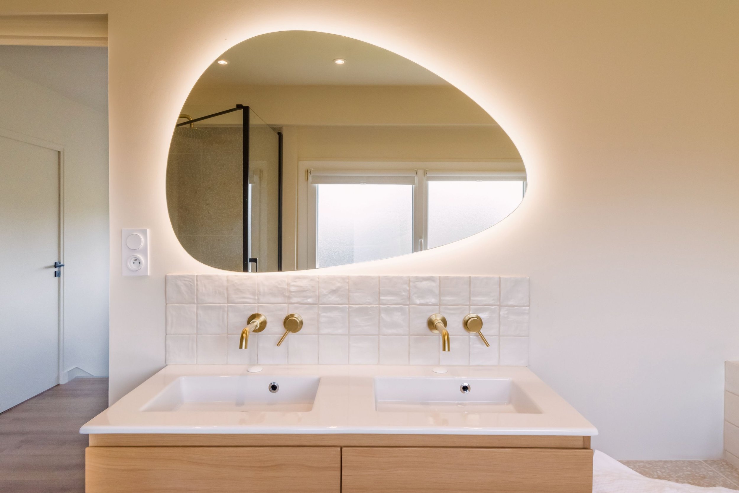 Un lavabo avec deux robinets en or et un miroir oval dans une salle de bain moderne. Fenêtre en arrière-plan avec vue partielle au-dessus du miroir.