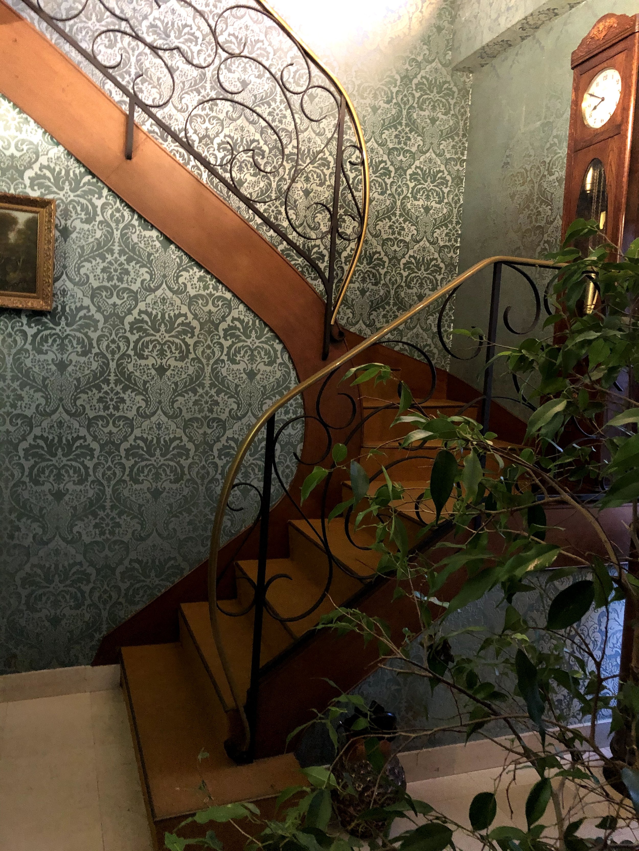 Escalier en bois avec une clôture en métal ornementale, decoré avec un mur tapissé d'un motif damassé en fond, une horloge en bois antique et une plante verte à côté.