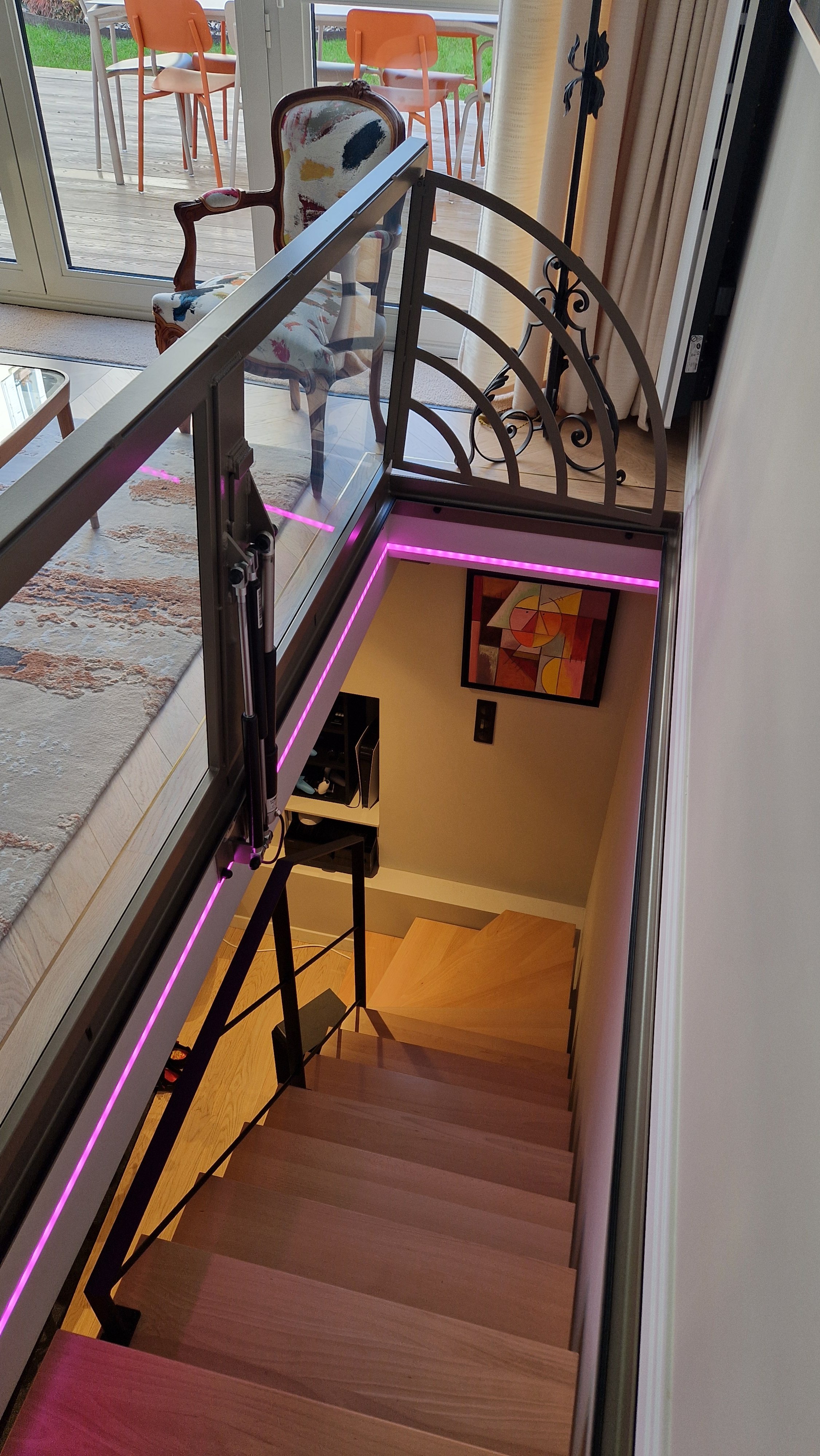 Escalier intérieur en bois avec éclairage LED rose, vue de haut, en face d'une salle de séjour avec une chaise vintage, une pièce d'art moderne sur le mur et une porte-fenêtre donnant sur un balcon avec des meubles orange.