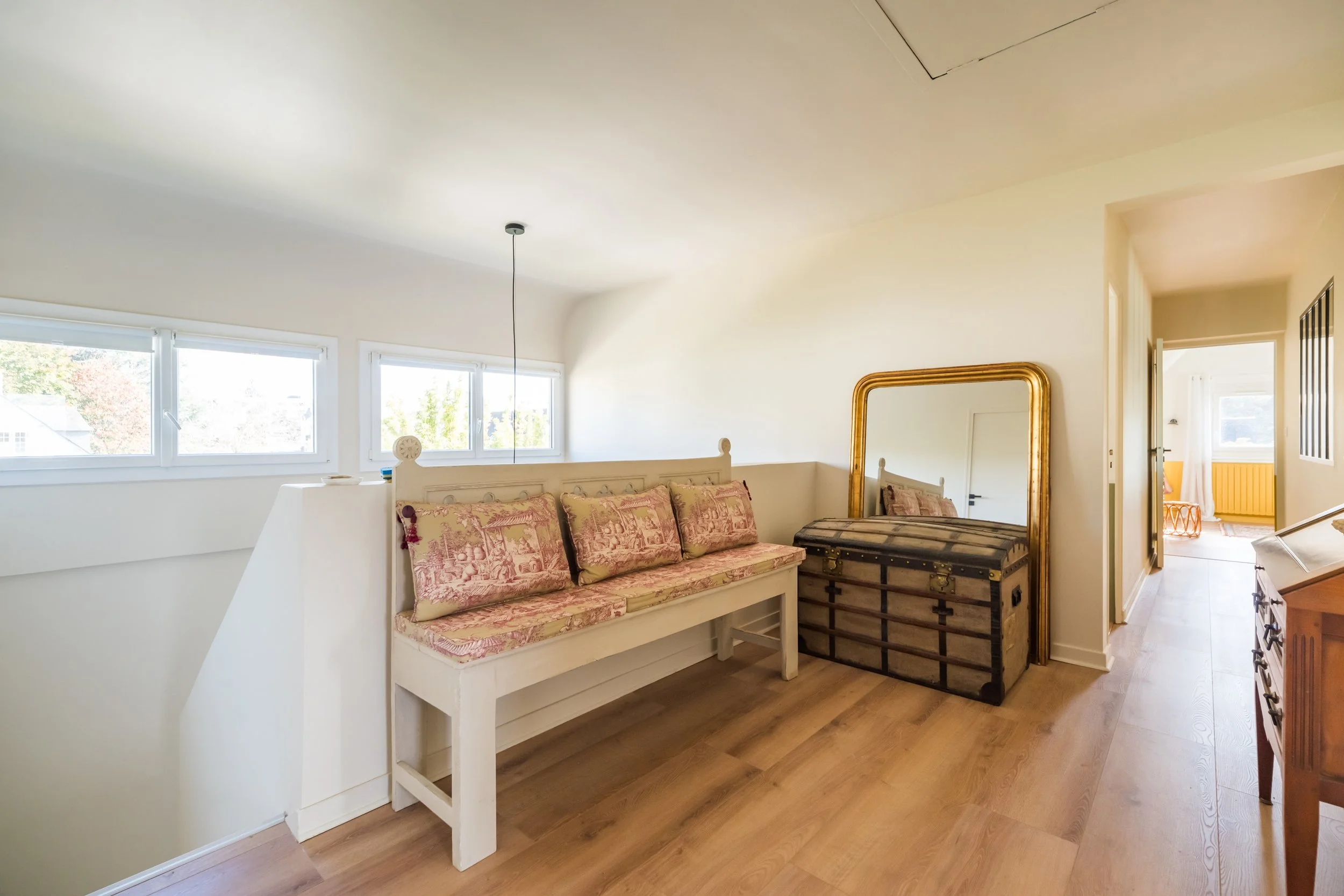 Une chambre lumineuse avec un lit en bois blanc, des coussins rose et or, une vieille malle en bois, un grand miroir à cadre doré et un mur blanc avec deux fenêtres.