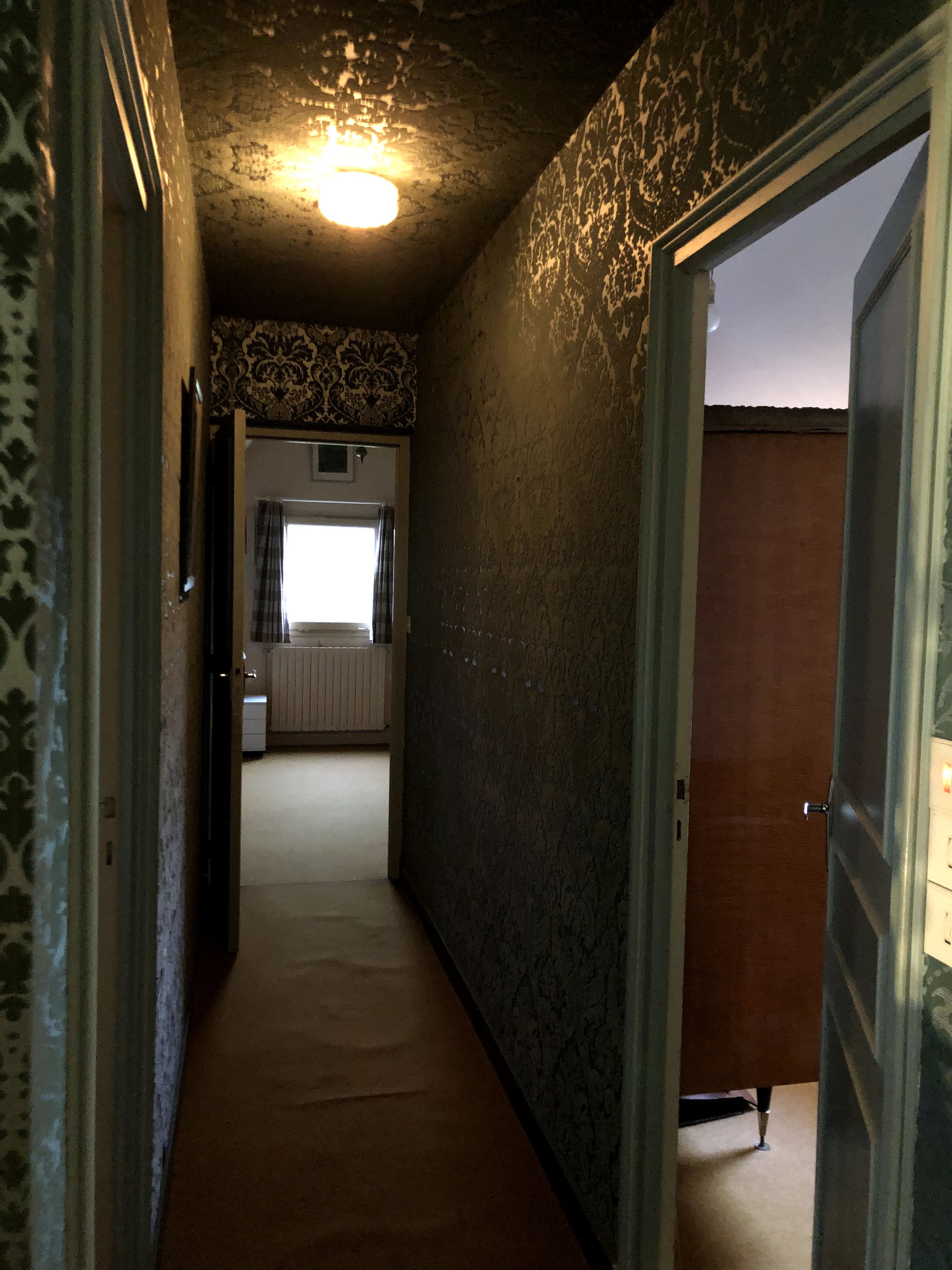 Un couloir dans une maison avec des murs recouverts de papier peint à motifs foncés, une lumière au plafond, une porte ouverte sur une pièce avec un radiateur, une fenêtre avec des rideaux et un lit visible en arrière-plan.
