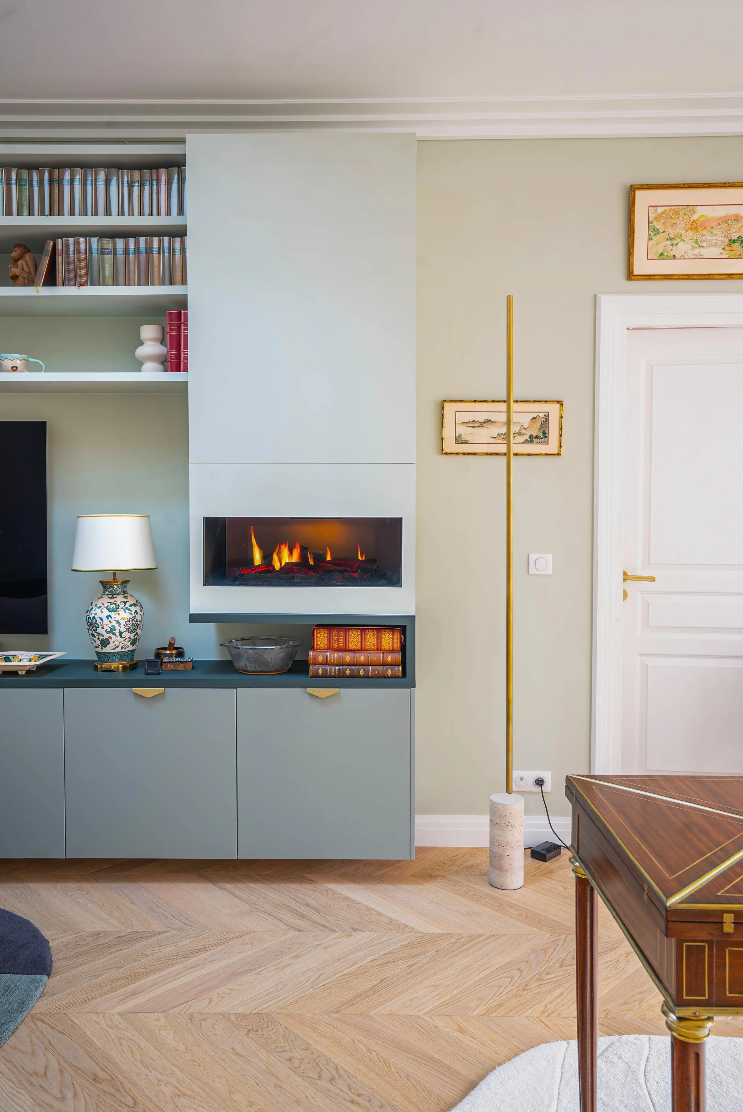 Intérieur d'un salon avec un meuble bleu-gris, une cheminée à feu électrique, des étagères remplies de livres, une lampe blanche avec base en céramique, une œuvre d'art encadrée, un lampadaire jaune en céramique blanche, un parquet en bois clair, et 