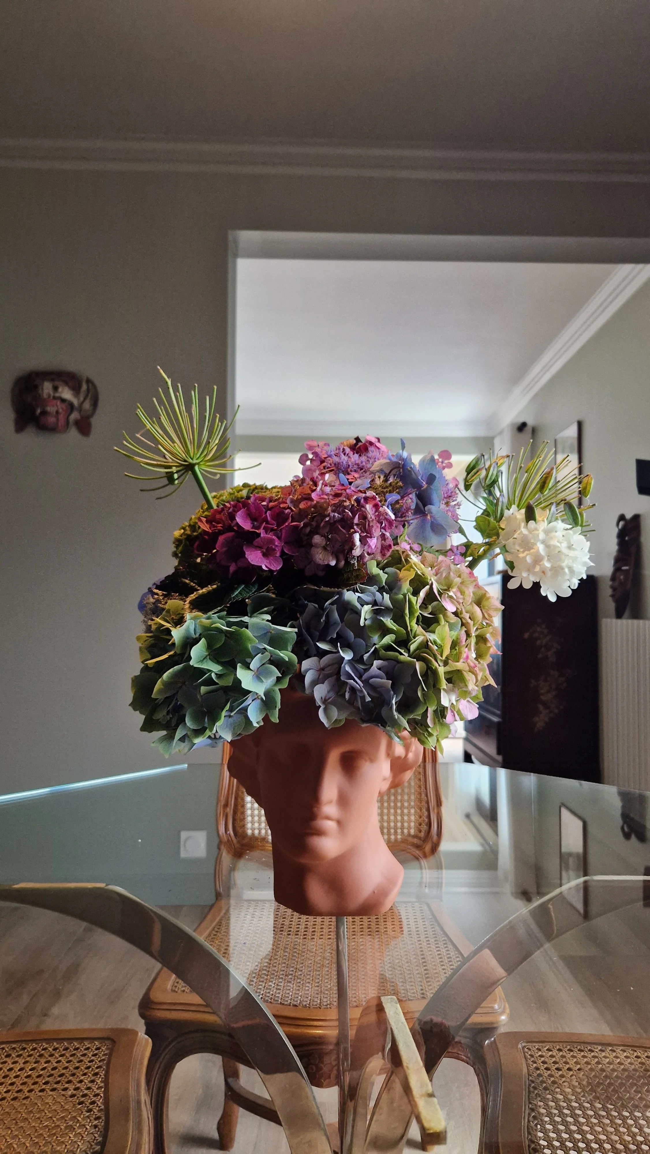 Un buste en céramique avec une tête humaine porte un arrangement floral composé de diverses fleurs, dont des hortensias, des roses et d'autres fleurs colorées.