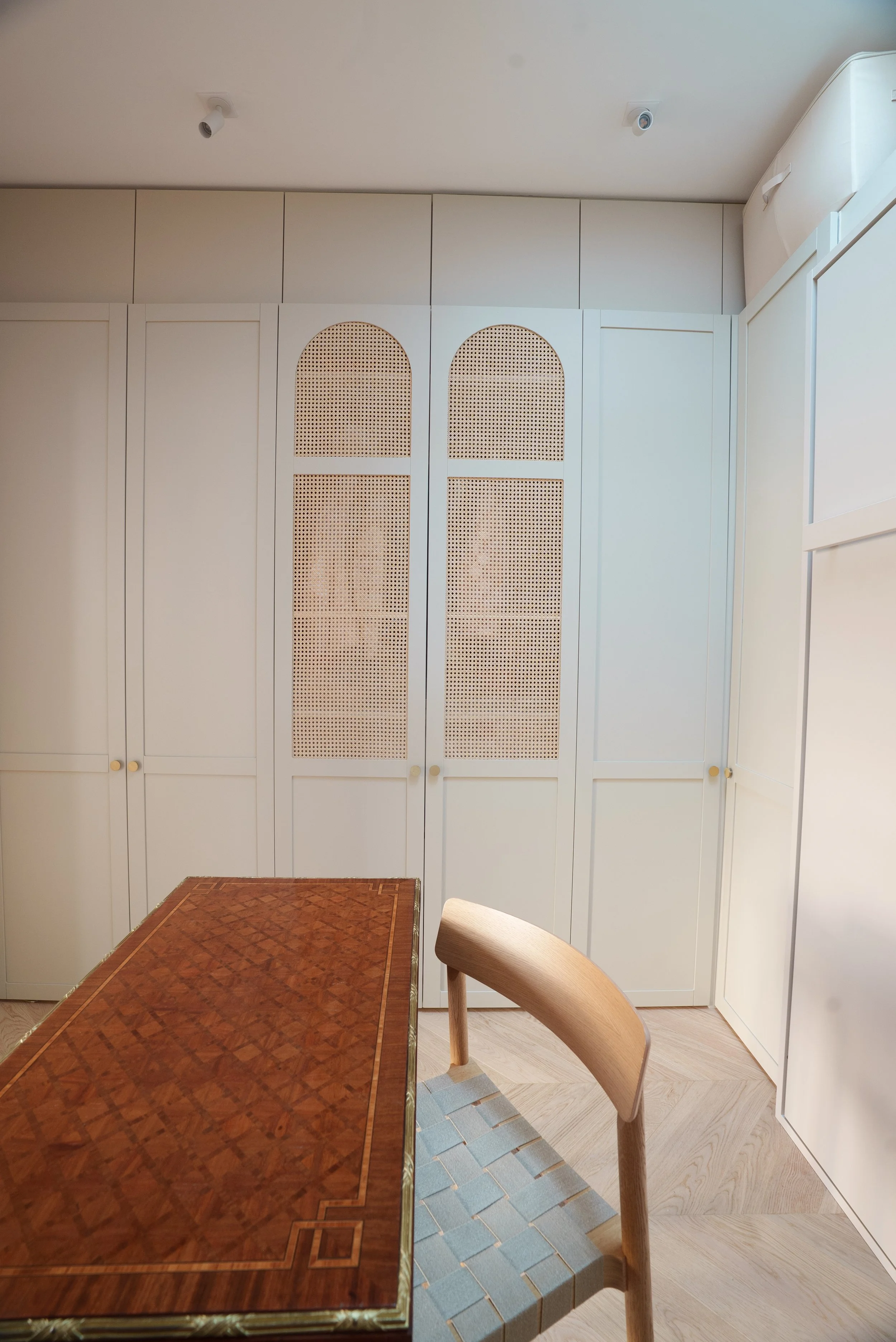 Une cuisine ou salle à manger avec meubles blancs, un meuble mural avec des portes à panneaux en bois perforé, une table en bois avec un motif en damier et une chaise en bois.