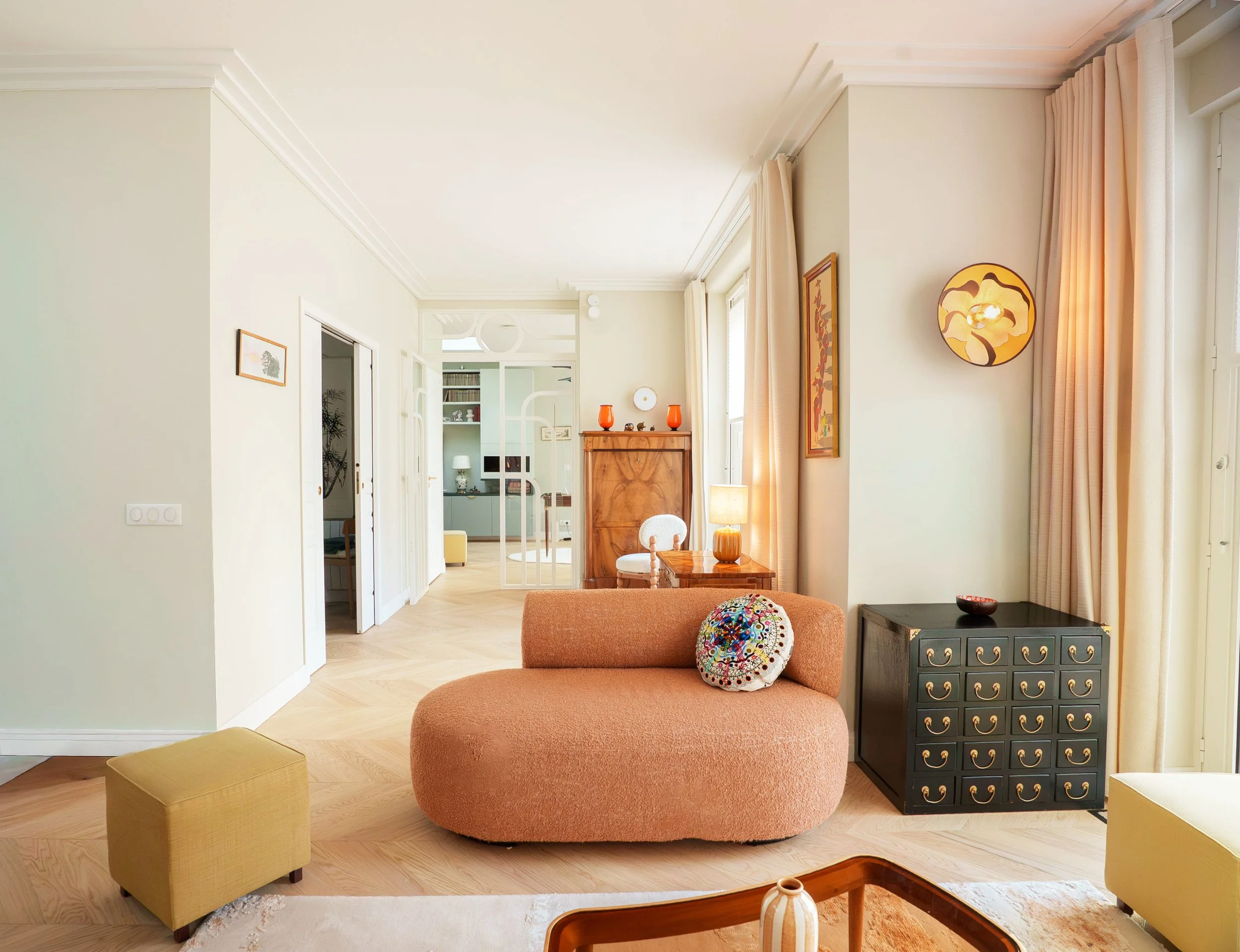 Intérieur d'un salon avec un canapé beige, un pouf jaune, une commode en bois, une lampe murale vintage, et des rideaux roses clairs.
