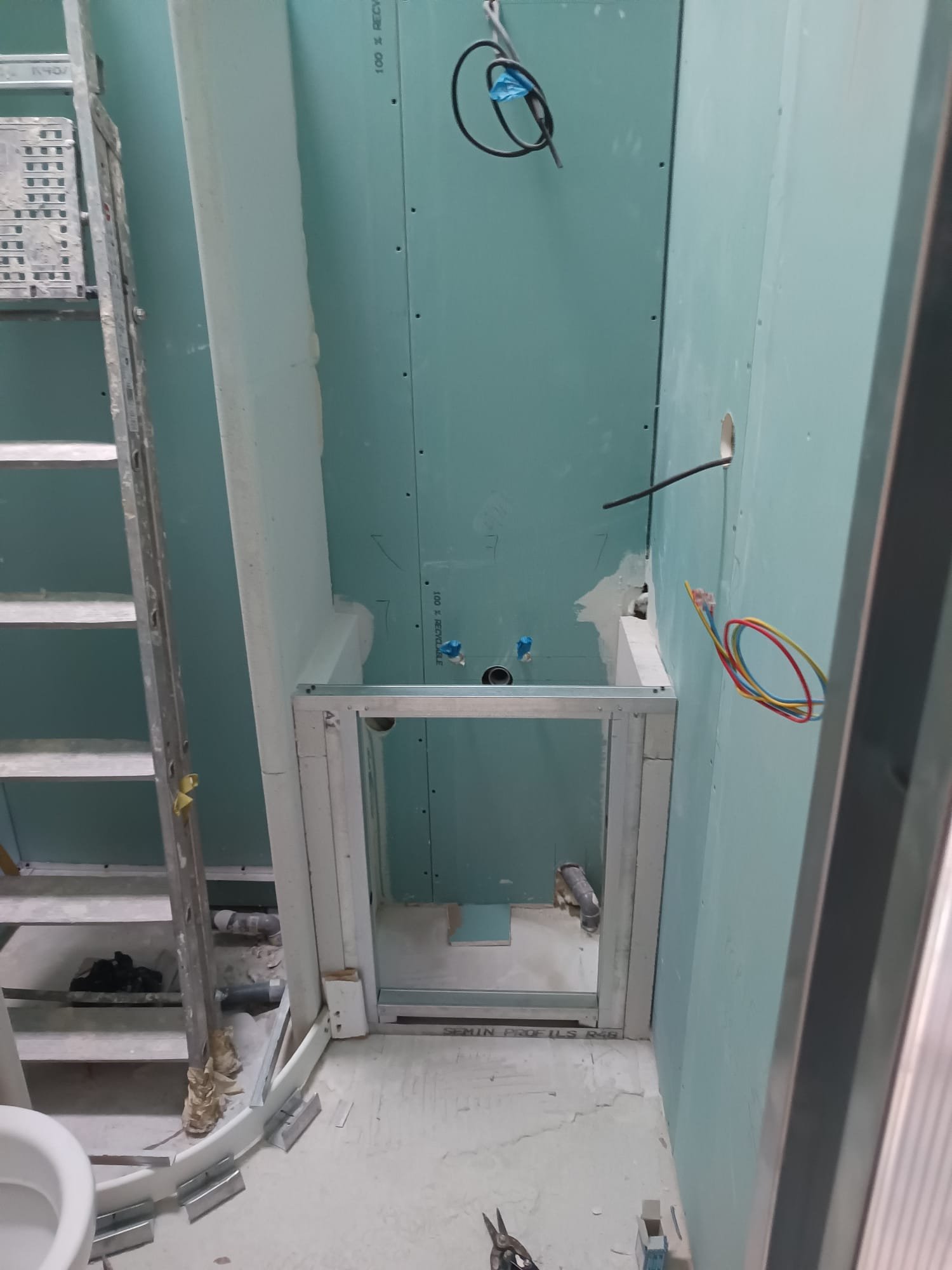 Salle de bain en cours de rénovation avec panneaux de cloison sèche, câbles électriques, tuyaux et équipements non installés, notamment un lavabo.