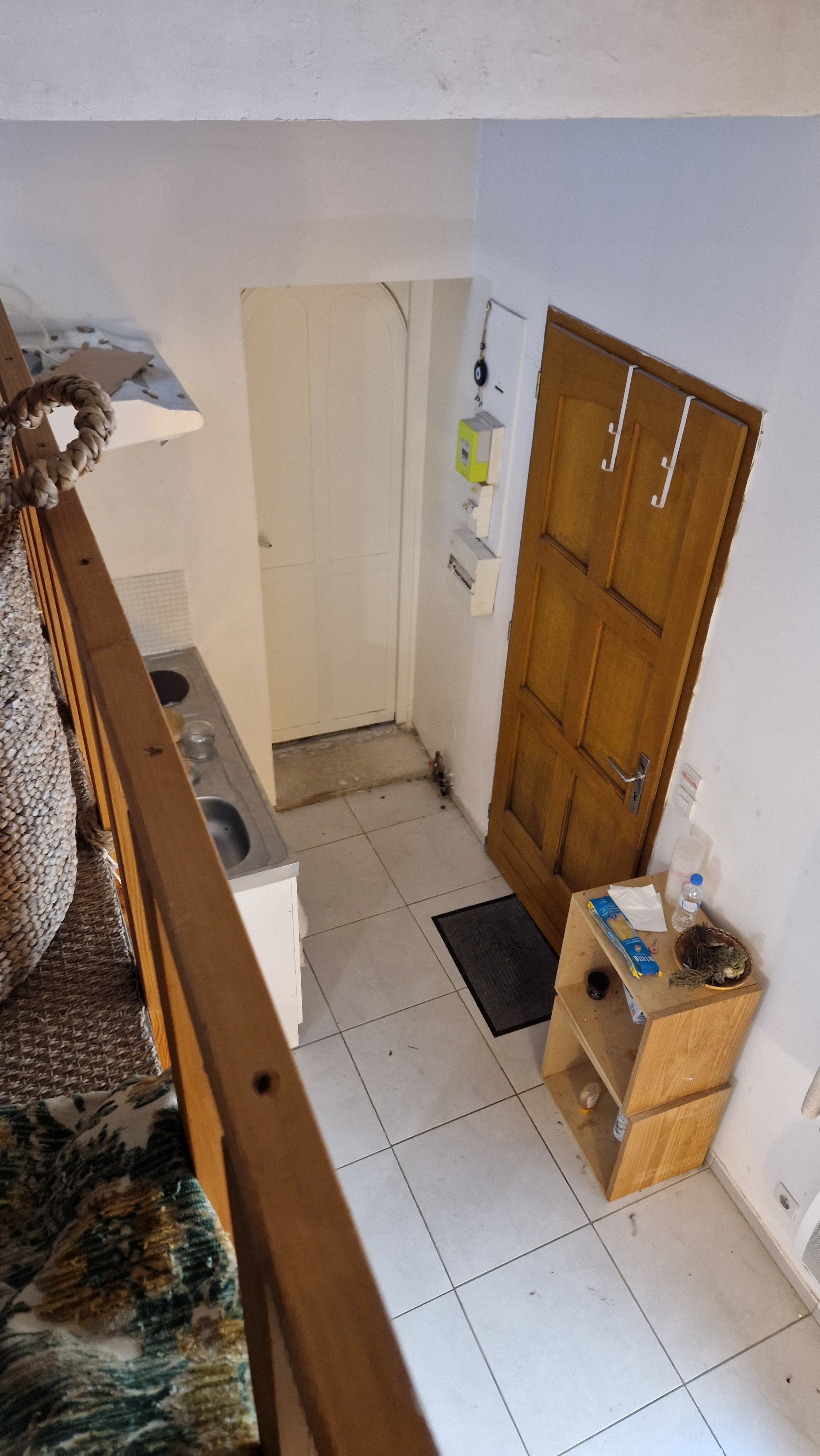 Intérieur d'une petite pièce, probablement un couloir ou une entrée, avec une porte en bois, une petite étagère en bois, un tapis noir, un évier en zone cuisine, quelques objets, cartes et bouteilles. Murs blancs et sol en carreaux.