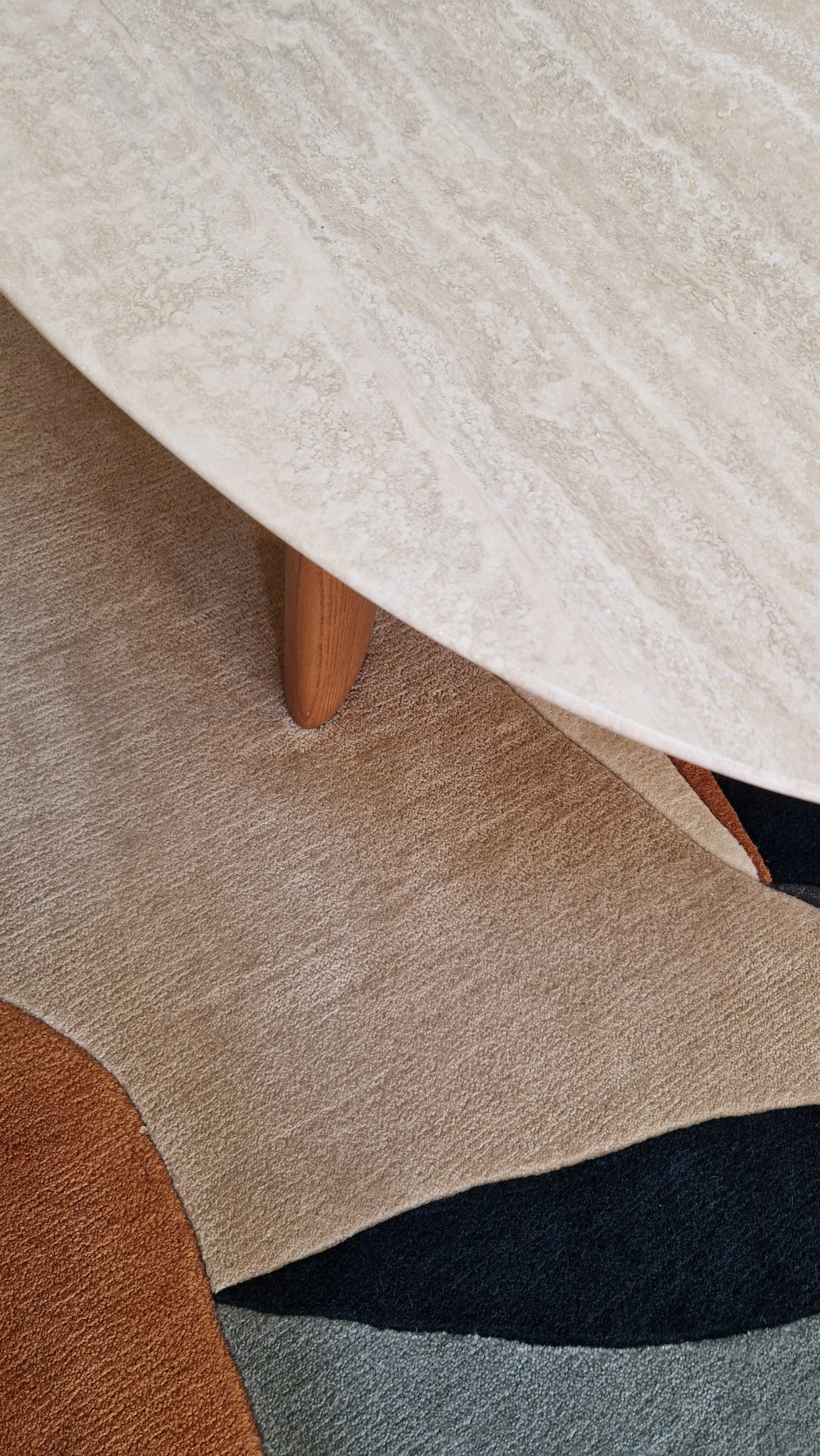 Table en pierre beige avec une jambe en bois clair, posée sur un tapis beige. Plusieurs tapis de différentes couleurs et textures sont visibles en dessous.
