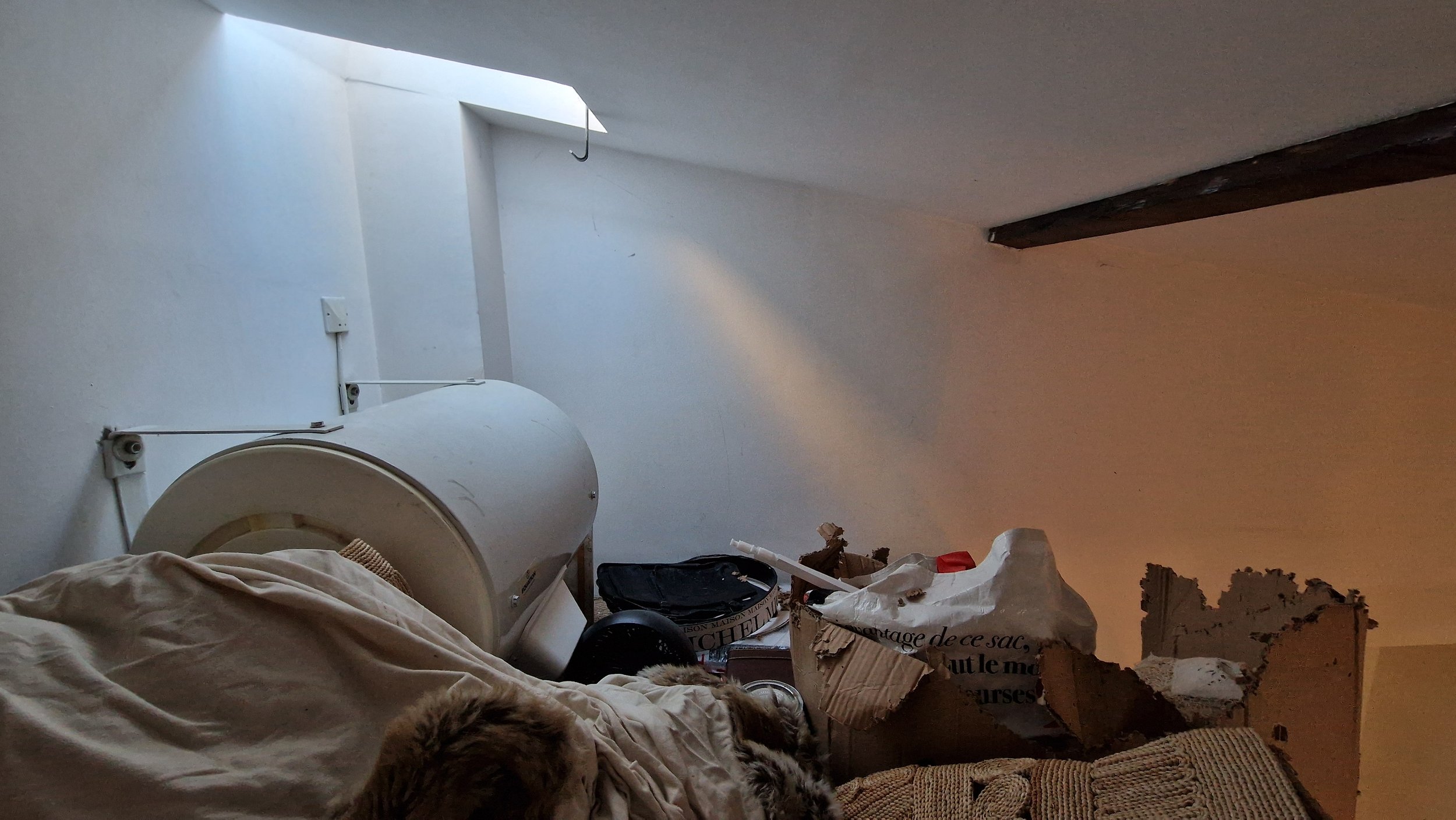 Chambre en désordre avec un lit, un radiateur d'antenne, des cartons, des sacs et des débris.
