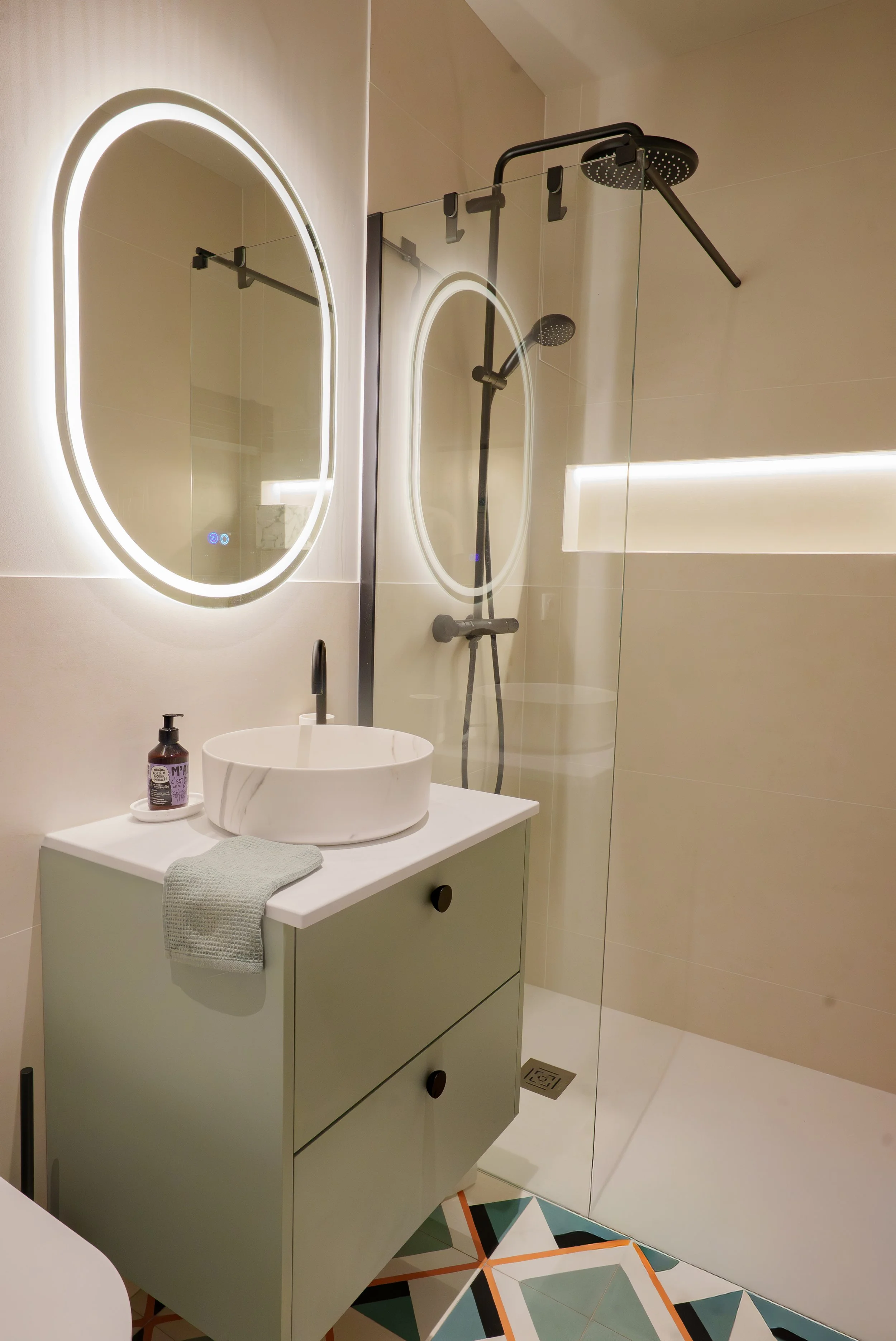 Une salle de bain moderne avec un miroir rond éclairé, un lavabo blanc à dessus rond sur une armoire verte clair, une douche avec paroi en verre et une douchette noire au plafond, un tapis gris et un sol avec des carreaux géométriques colorés.