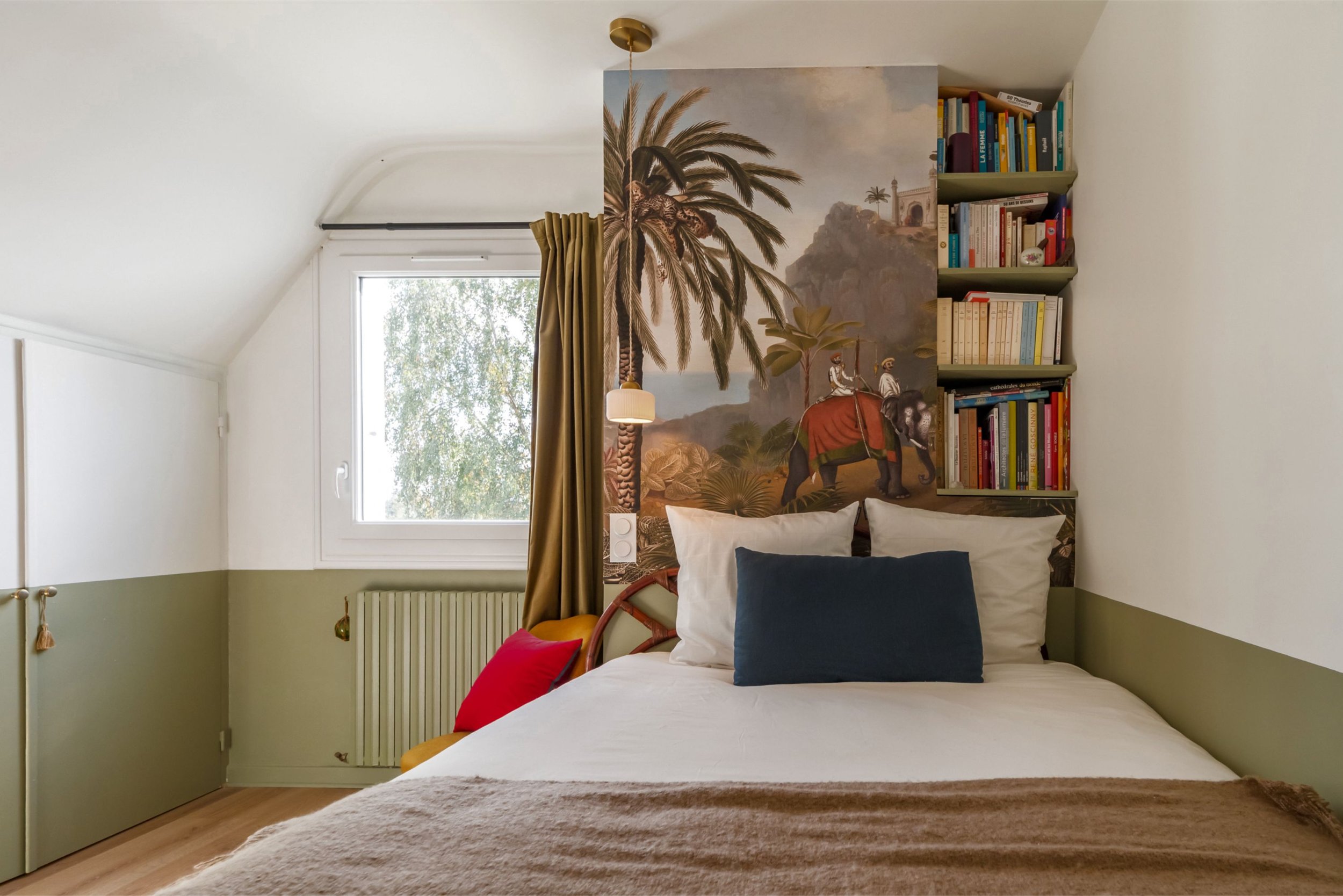 Une chambre avec un mur peint d'une scène de désert tropical avec des palmiers, deux personnes à dos assises sur un éléphant, une grande fenêtre laissant entrer la lumière naturelle, un lit avec plusieurs coussins.