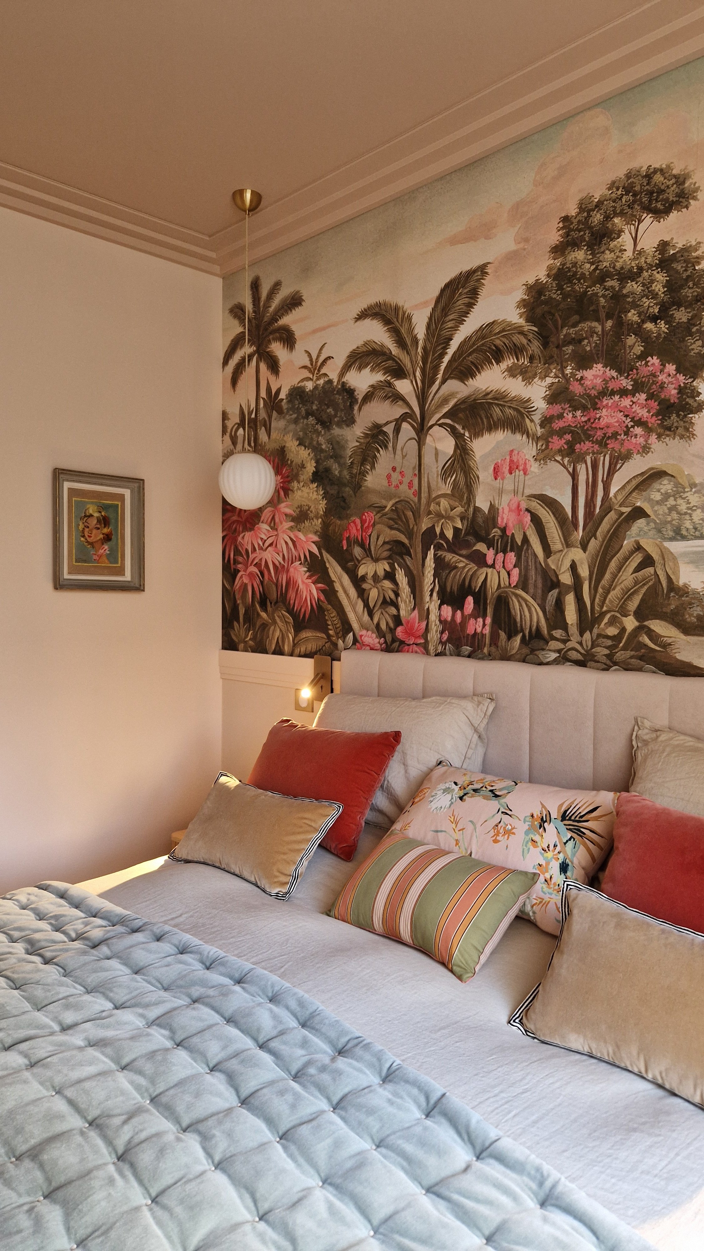 Chambre avec un mur en fresque représentant un paysage tropical avec des palmiers et des fleurs roses, un lit avec plusieurs coussins de différentes couleurs et un cadre avec une peinture sur le mur latéral.
