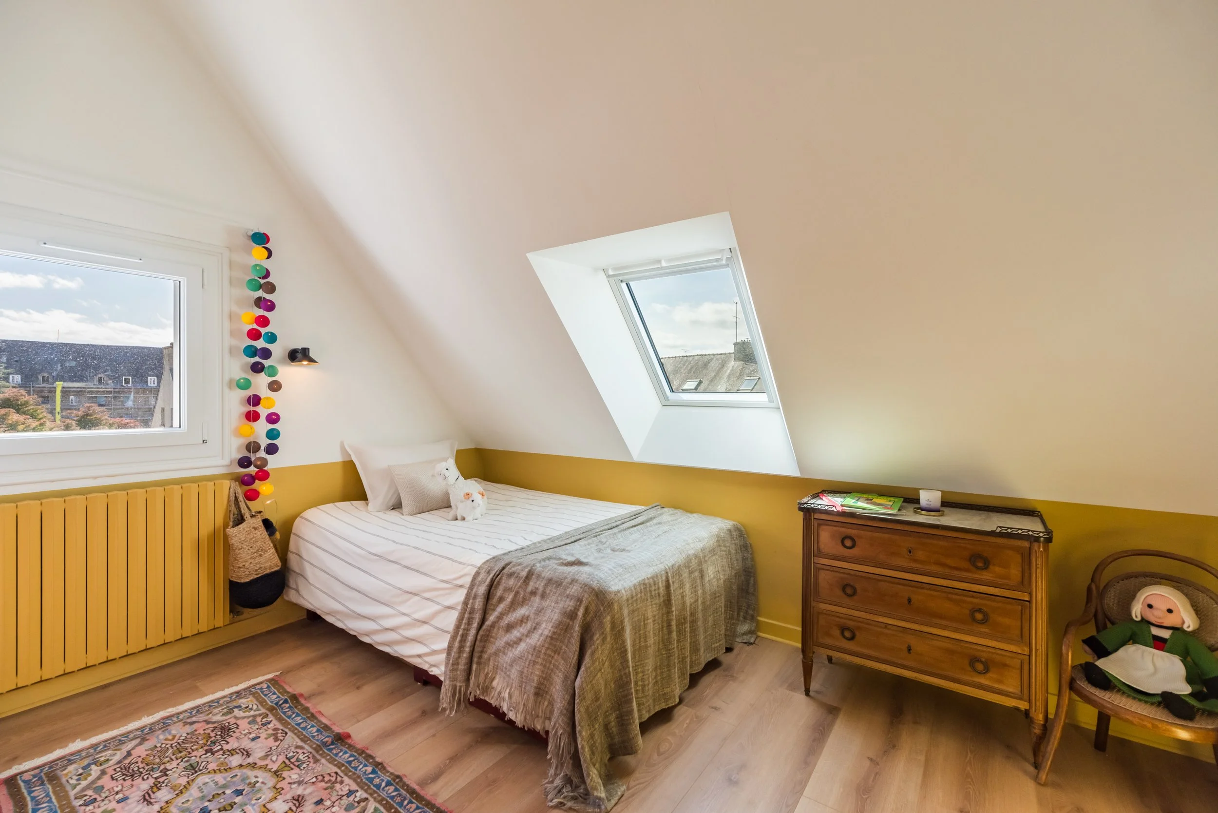 Chambre avec lit double, fenêtres en velux, décor lumineux et coloré, avec une étagère en haut avec des boules multicolores et un petit bureau en bois.