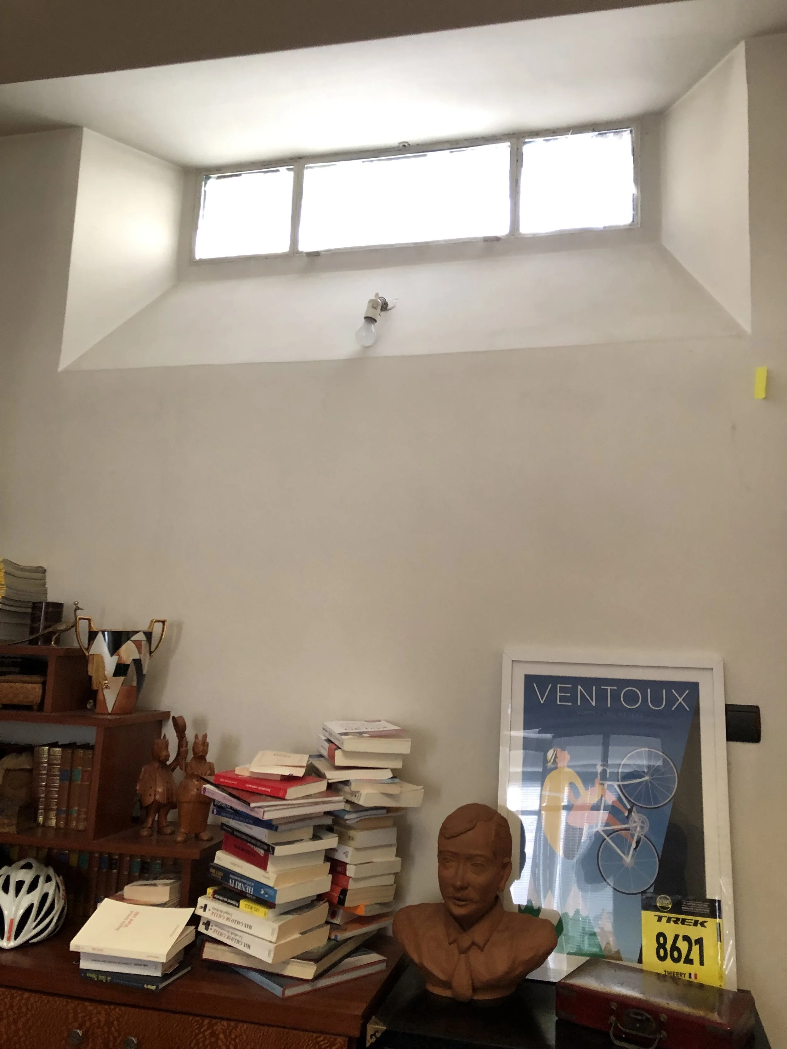 Une étagère en bois avec plusieurs livres empilés, une sculpture en bronze d'un homme à col roulé, un casque de vélo blanc avec noir, une affiche avec le texte 'VENTOUX' devant une fenêtre, une médaille de course 'TREK 8621' avec la mention 'Thierry'