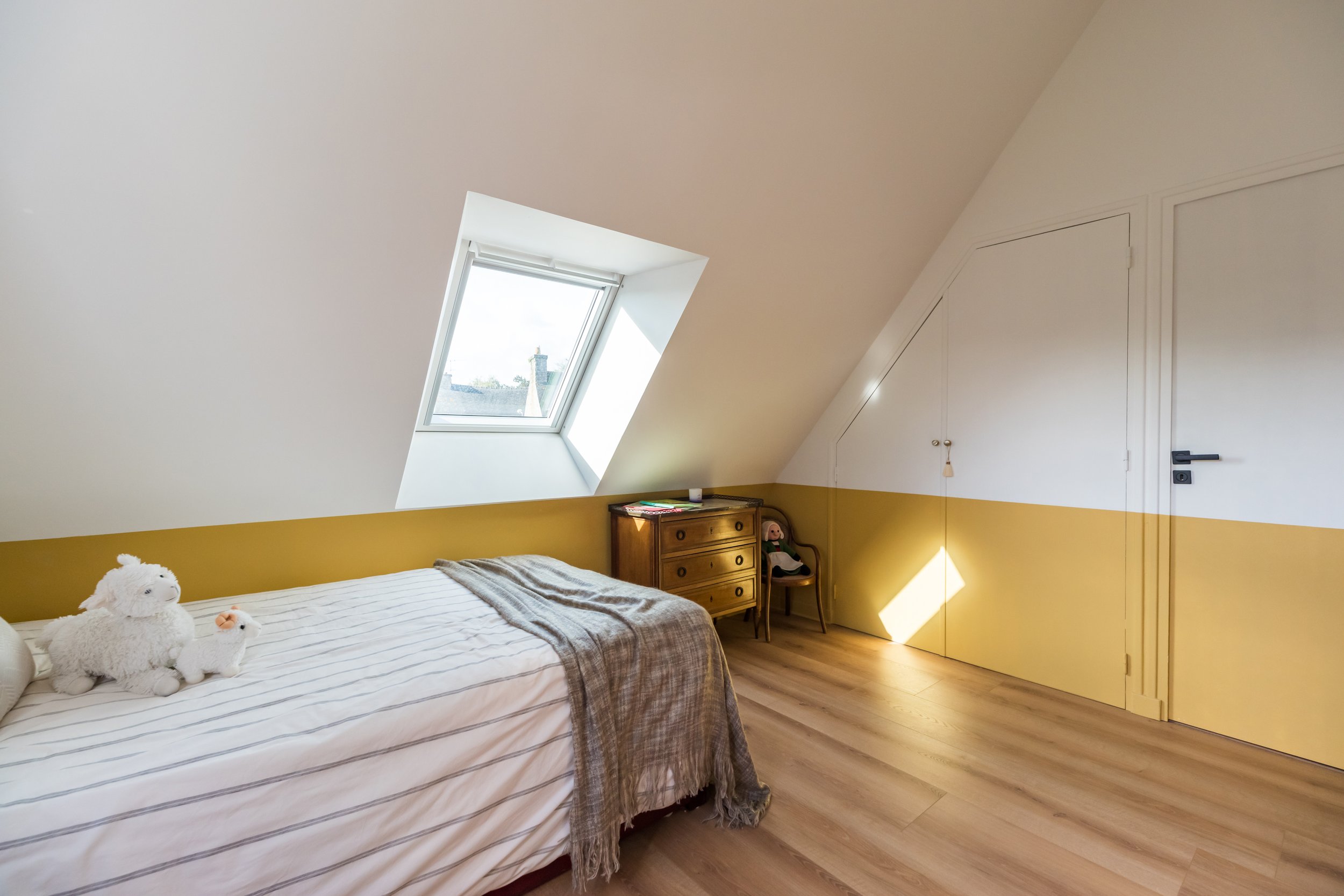 Chambre avec un lit, deux ours en peluche, un bureau en bois, un fauteuil, une porte, un coffre avec des livres, et un plafond avec une lucarne offrant une vue sur le ciel.