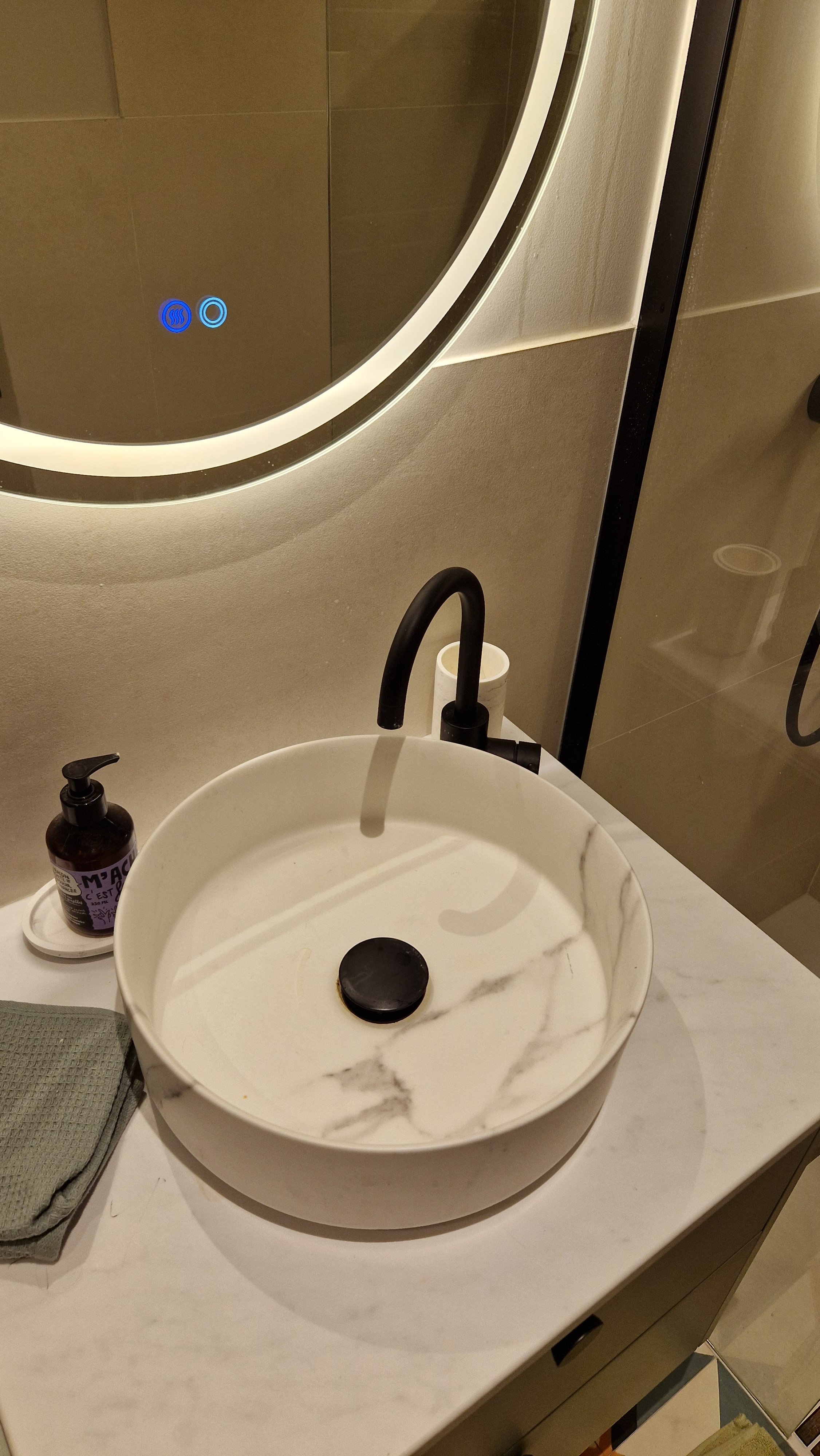 Un lavabo rond en marbre blanc avec des veines grises, une robinetterie noire, un miroir rond avec éclairage LED, un savon liquéfié dans un distributeur, un petit pot blanc, et une serviette grise à côté.