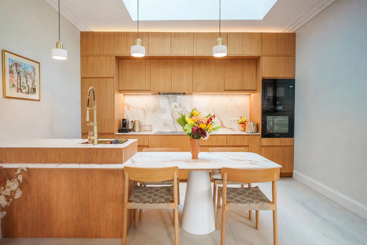Cuisine moderne avec mobilier en bois clair, plan de travail en marbre blanc, étagères et électroménagers intégrés. Au centre, table ronde avec vase de fleurs, entourée de trois chaises en bois, sous un éclairage suspendu.