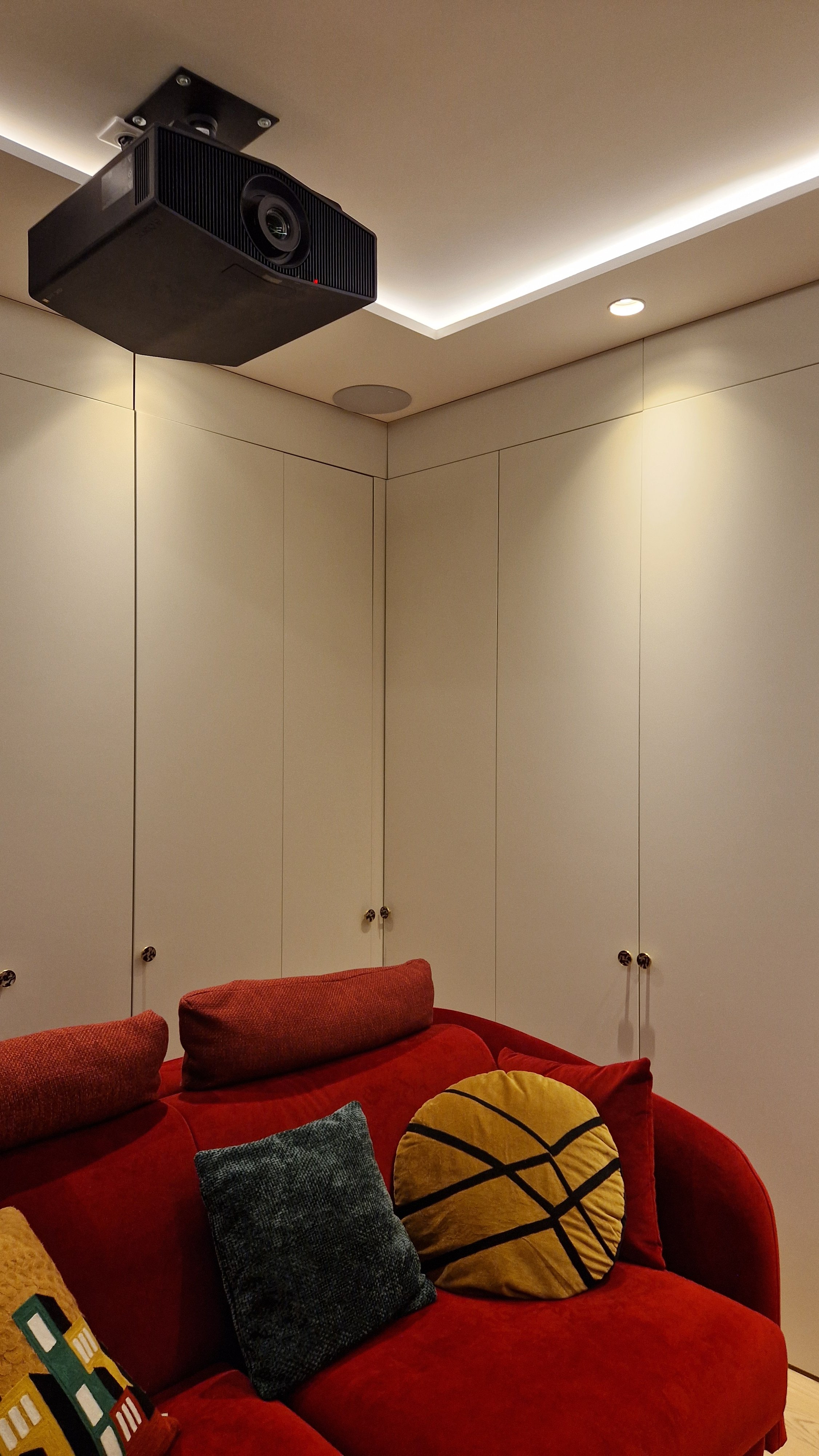 Salon avec un canapé rouge, plusieurs coussins colorés, et un mur blanc avec des armoires et un projecteur suspendu au plafond