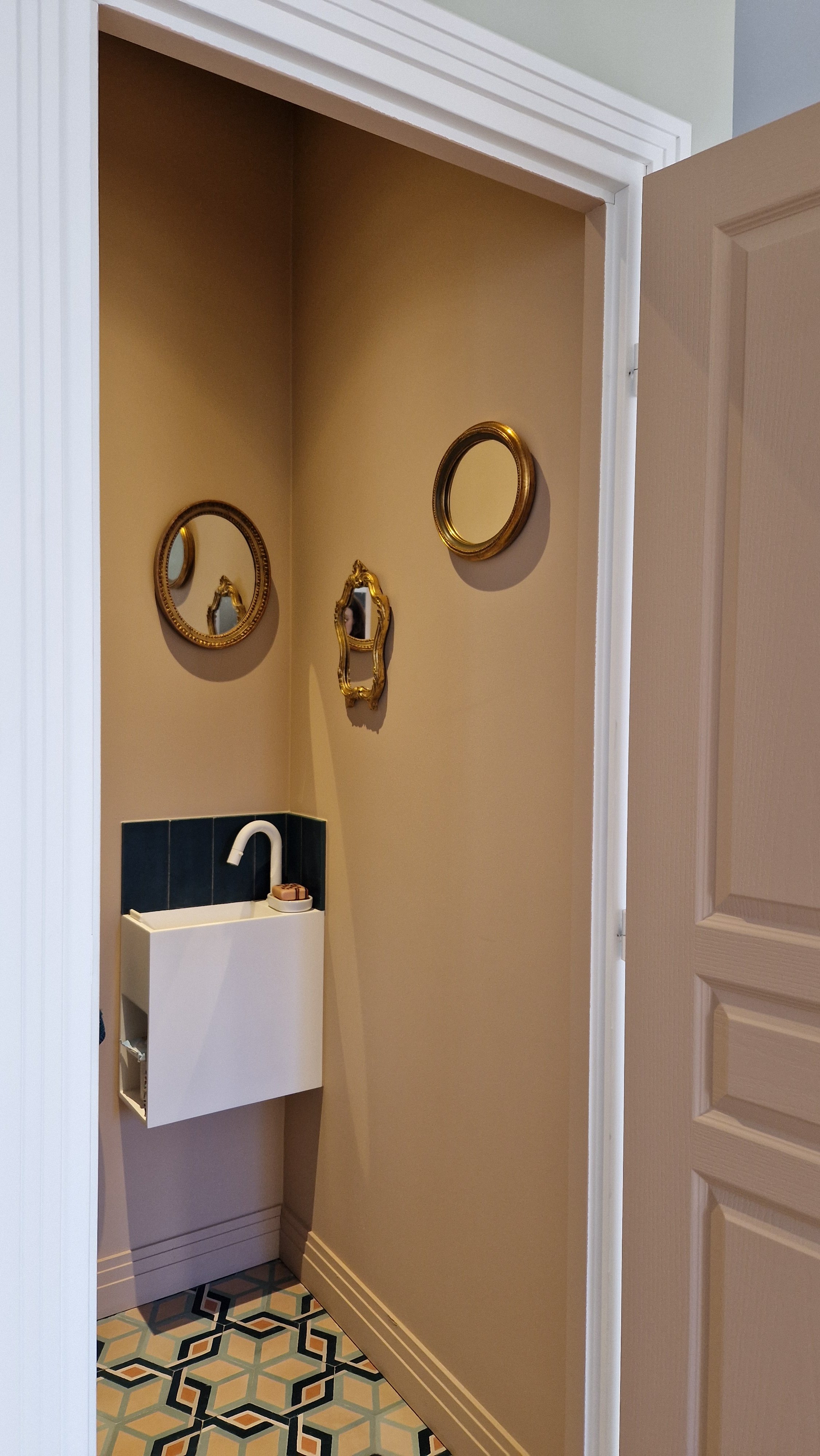 Petite pièce avec une étendue beige, des miroirs dorés au mur, un évier blanc avec un robinet blanc, un savon et un petit objet, séparée par une porte en bois clair, avec un sol à motifs géométriques colorés.