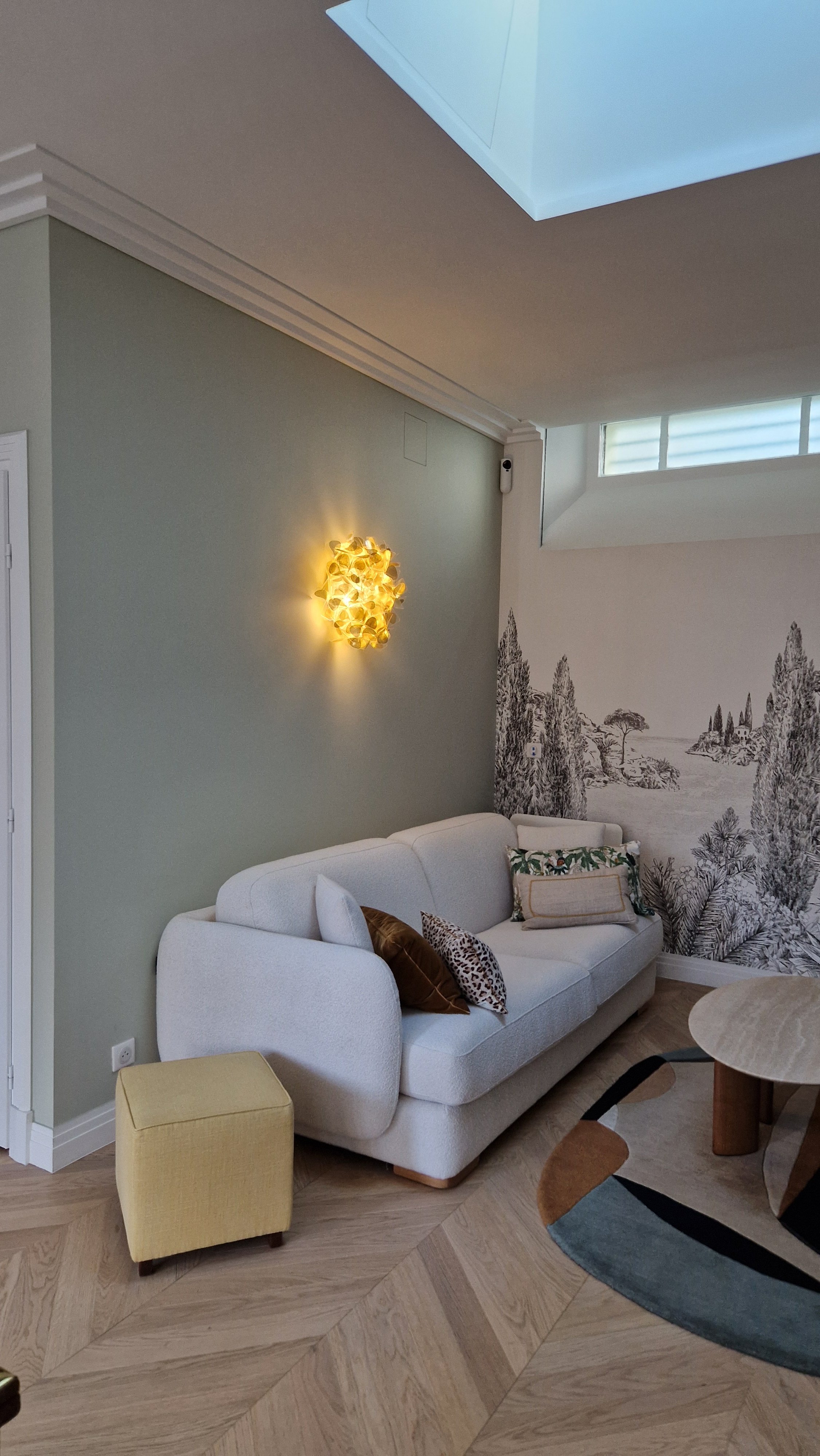 Intérieur d’un salon moderne avec un canapé blanc, des coussins, un tapis coloré, un mur avec une fresque et une suspension lumineuse