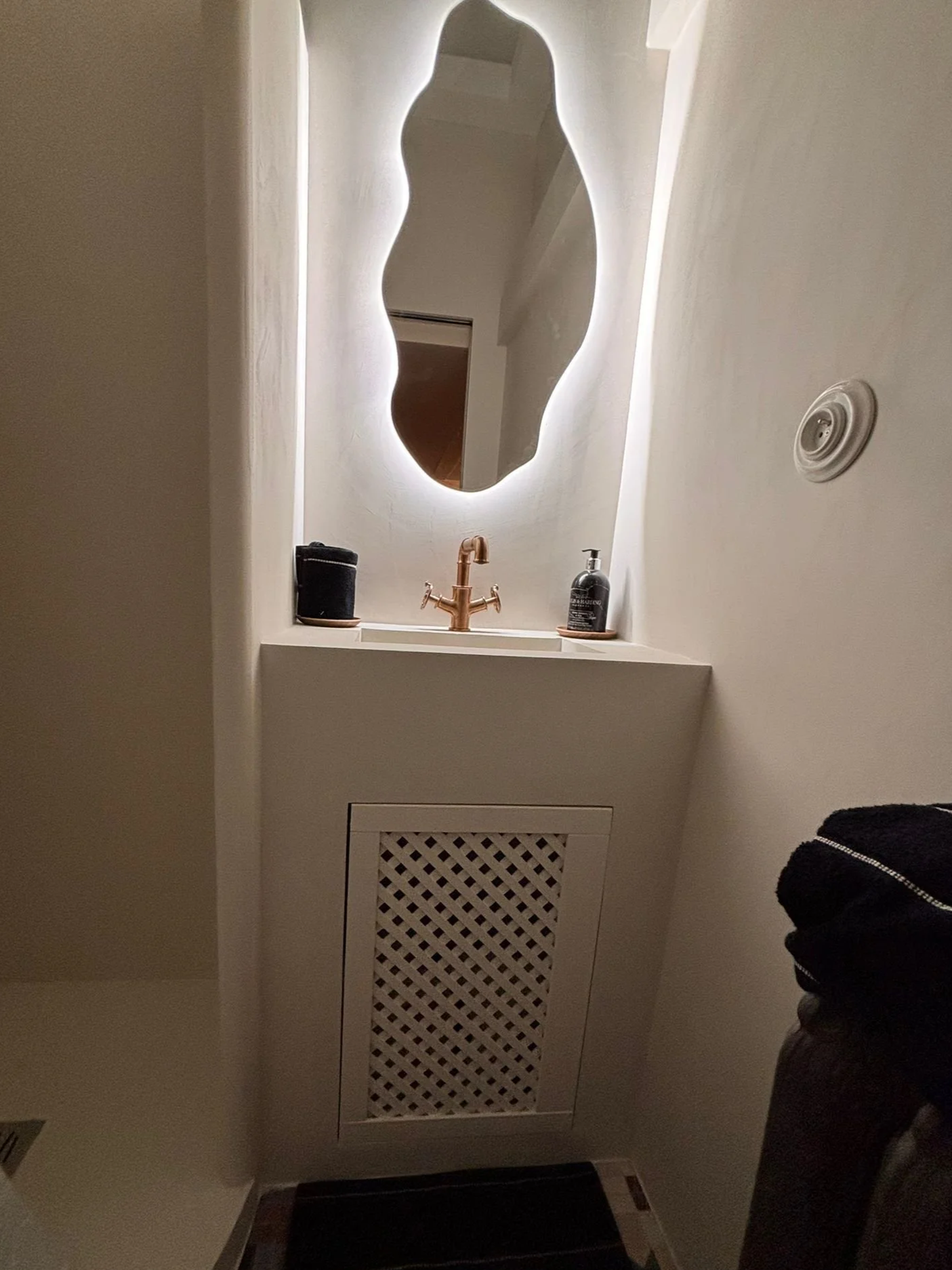 Miroir avec éclairage intégré au-dessus d'un lavabo en cuivre, avec un savon et un récipient noir sur le rebord, à côté d'une porte en bois et d'un radiateur en dessous, dans une pièce intérieure.