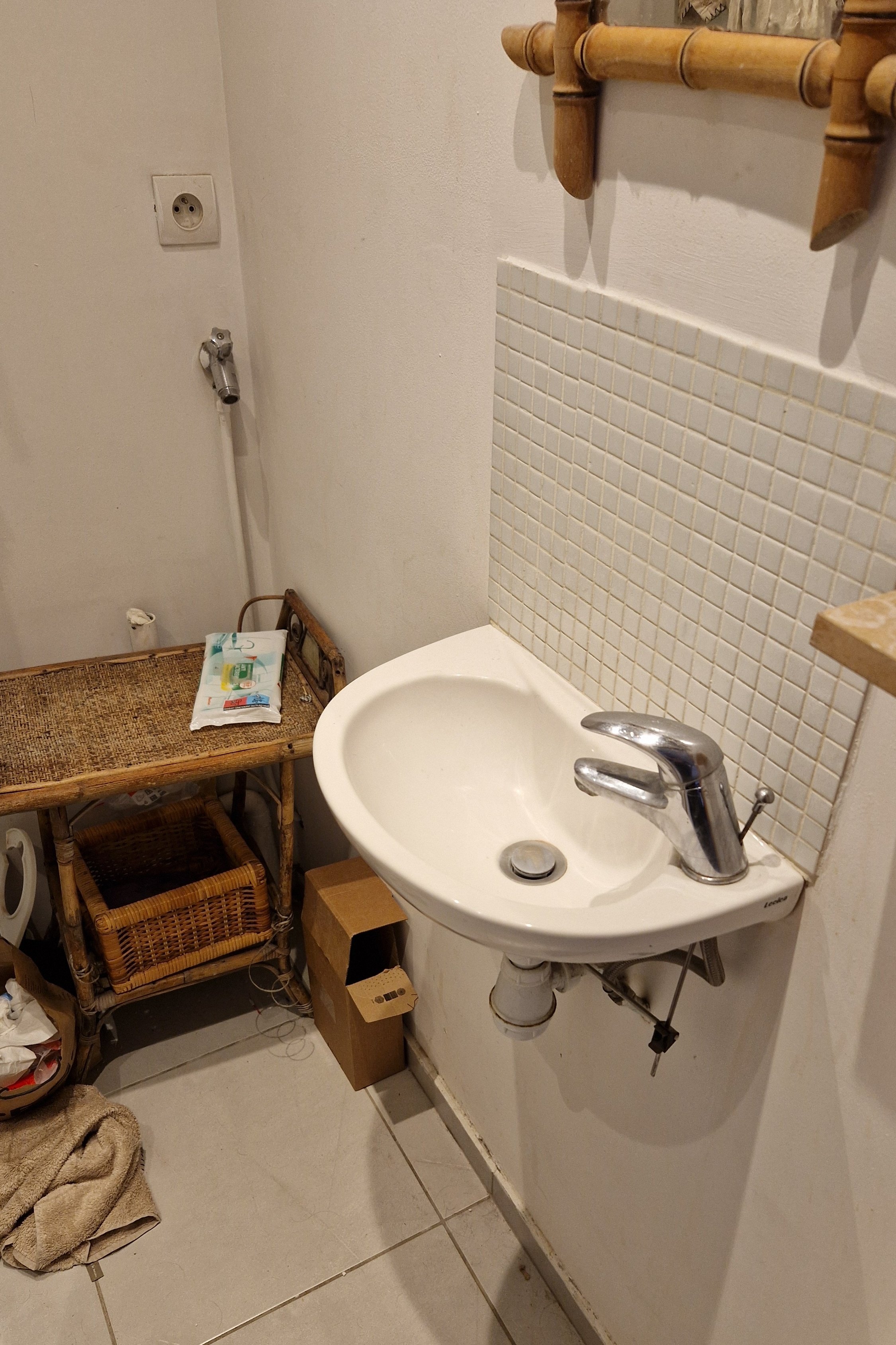 Photo d'une petite salle de bain désordonnée avec un lavabo blanc, un miroir en bois, un tabouret en bois avec un paquet de lingettes, une caisse en carton, un panier en rotin, des serviettes enroulées et des câbles électriques.