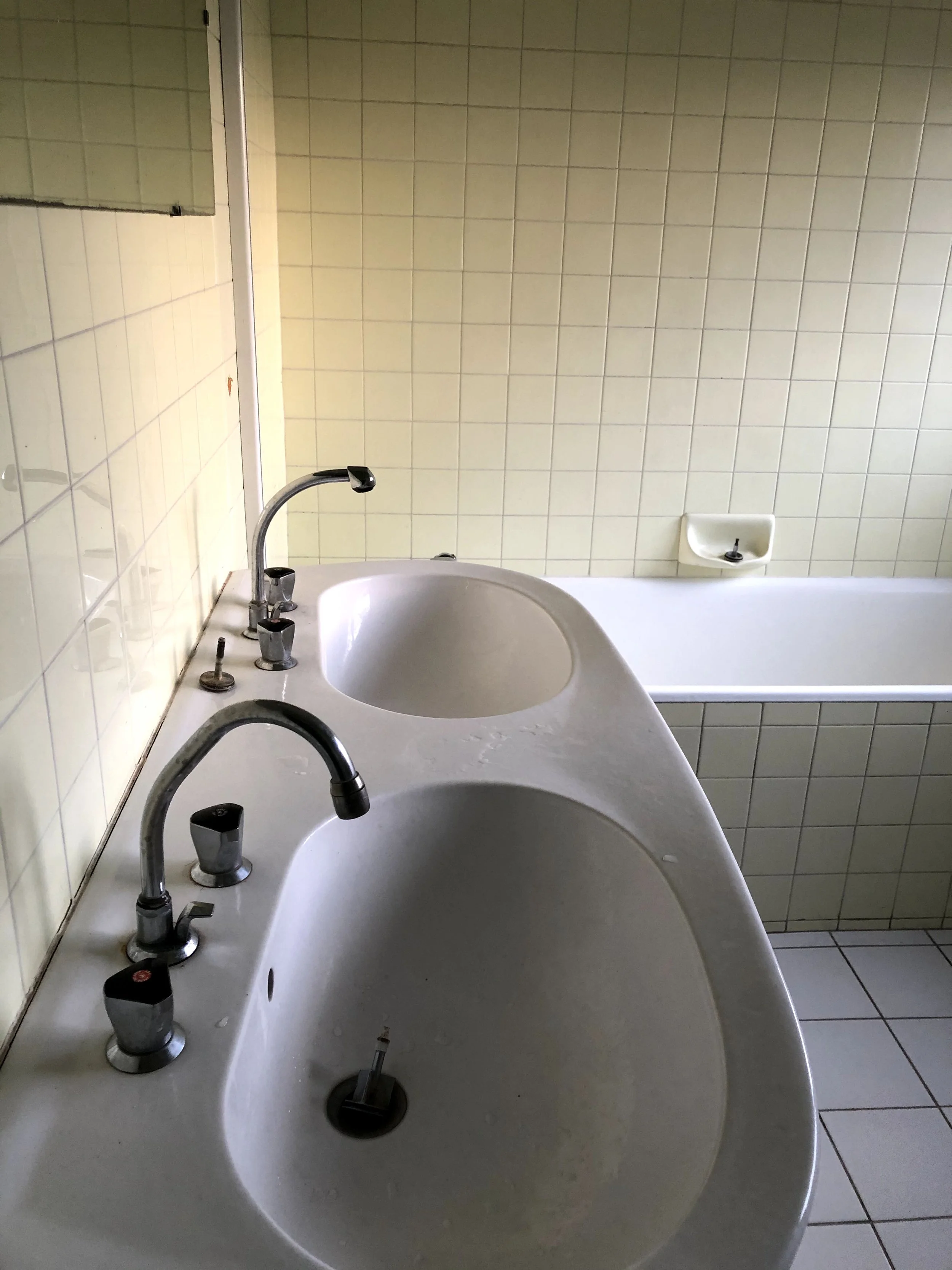 Vasque de salle de bain avec deux robinets et miroir au-dessus, mur carrelé de couleur beige.