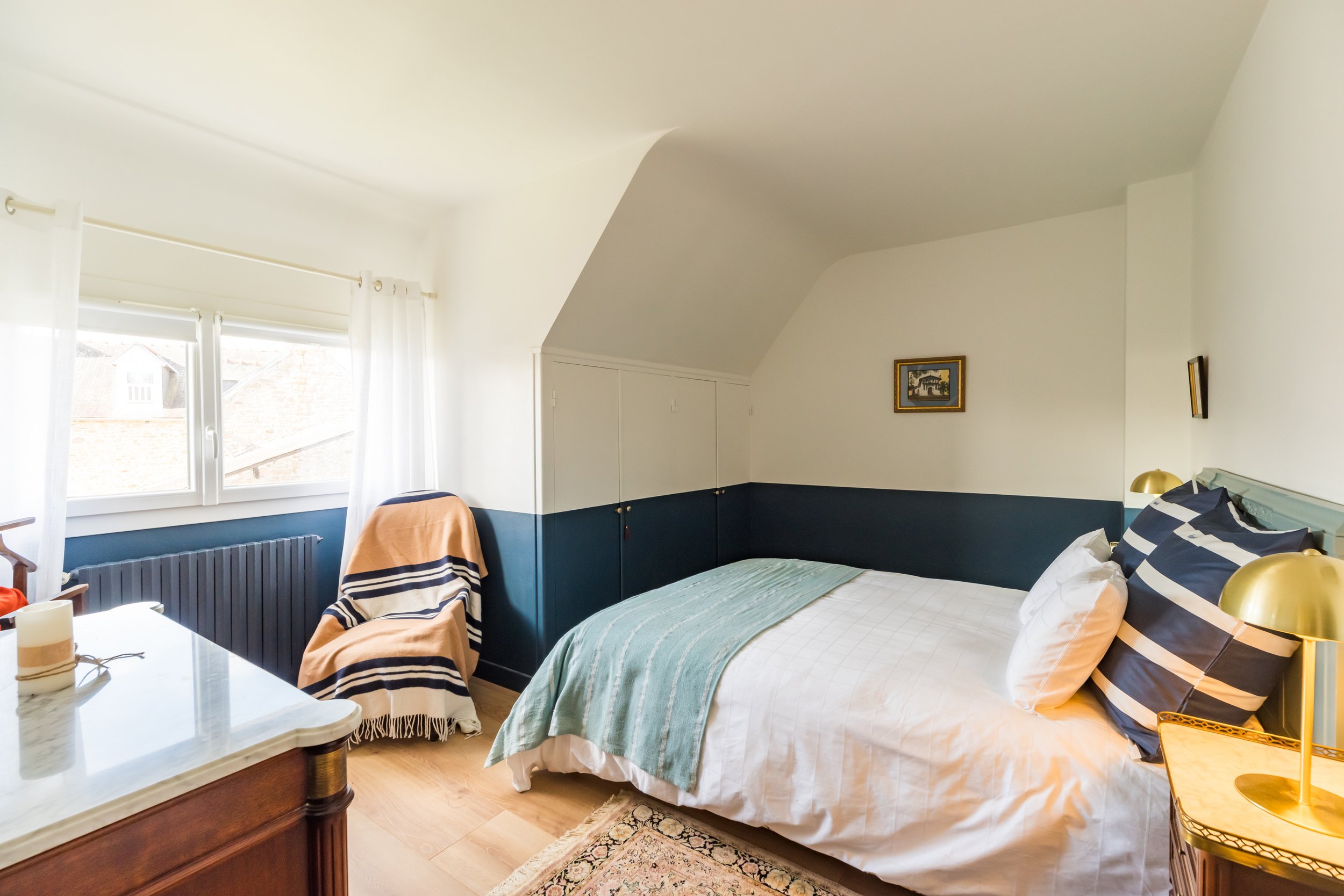 Chambre lumineuse avec un lit double, couvert de draps blancs et de coussins rayés. Fenêtres avec rideaux blancs laissant entrer la lumière. Mur blanc avec une décoration artistique. Meubles comprenant une tête de lit, une petite table dorée, une com