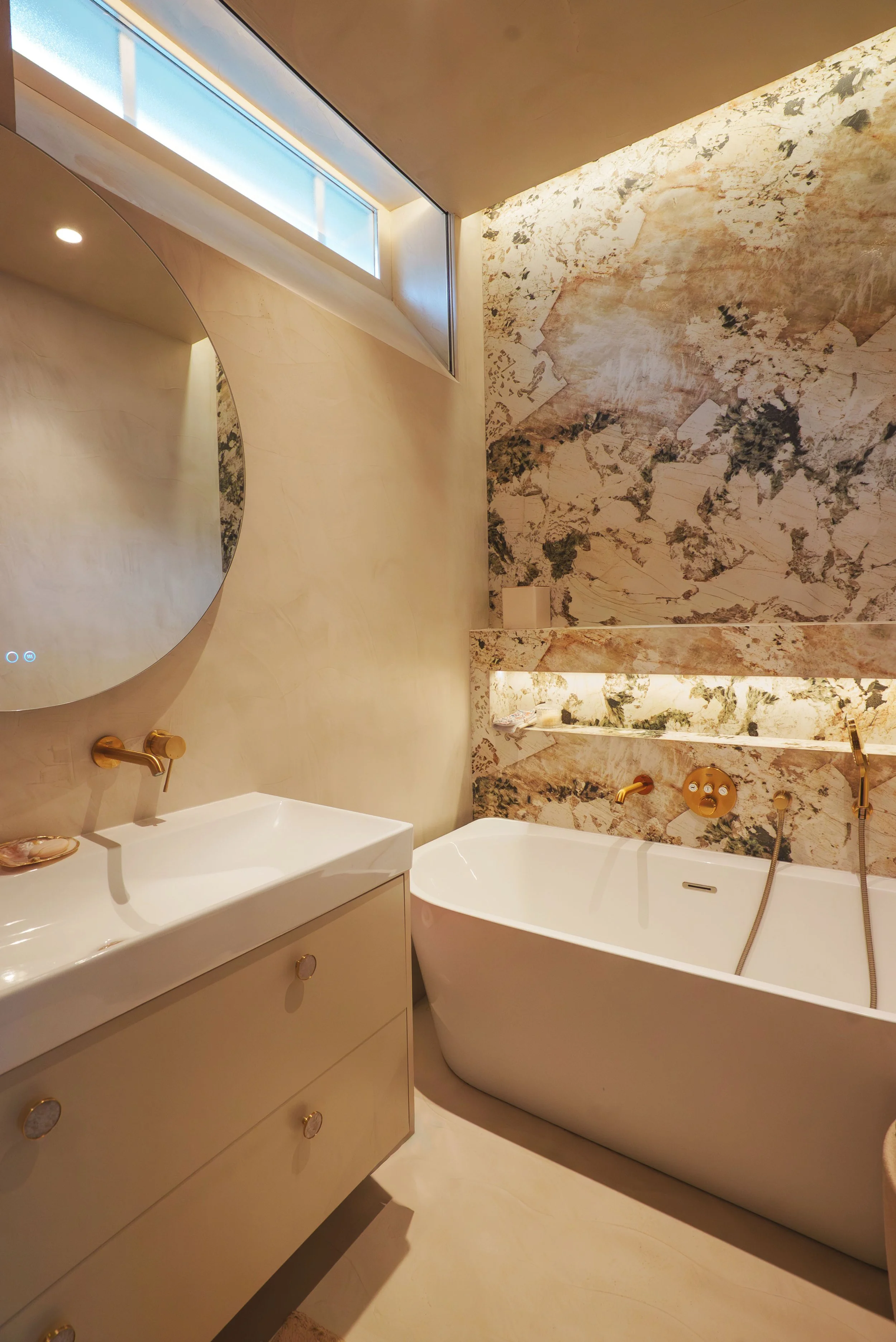 Une salle de bains élégante avec un lavabo, un miroir rond, une baignoire en coin, un mur en marbre et des robinets dorés.