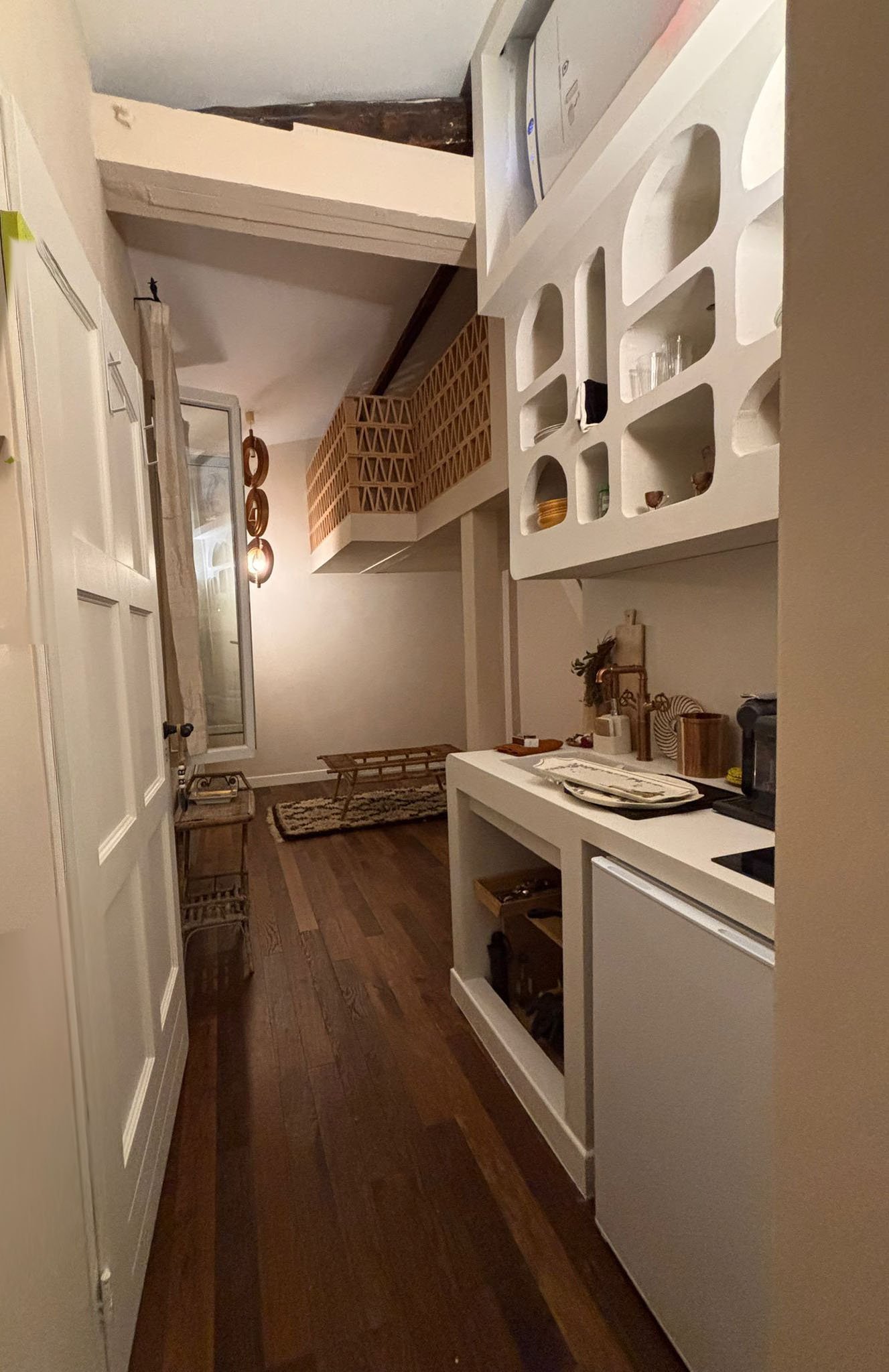 Intérieur d'un appartement avec cuisine équipée, meuble blanc avec niches, plan de travail, et un coin salon au fond avec tapis, meubles en bois, et un mur décoré de suspensions en bois.