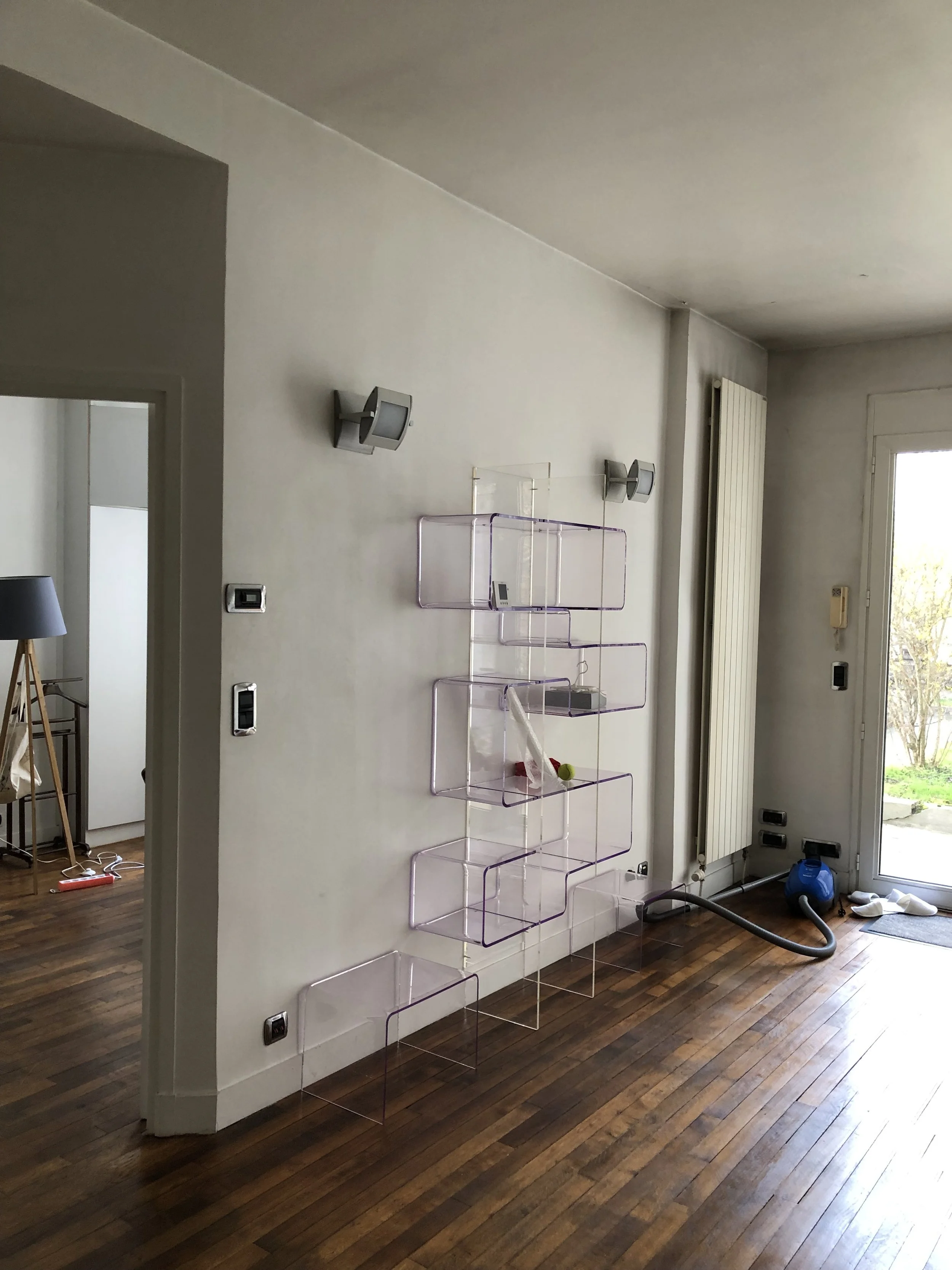 Intérieur d'une pièce avec un mur blanc, plusieurs étagères en plastique transparent violet, un radiateur blanc, une fenêtre avec vue sur un jardin, et un sol en bois.