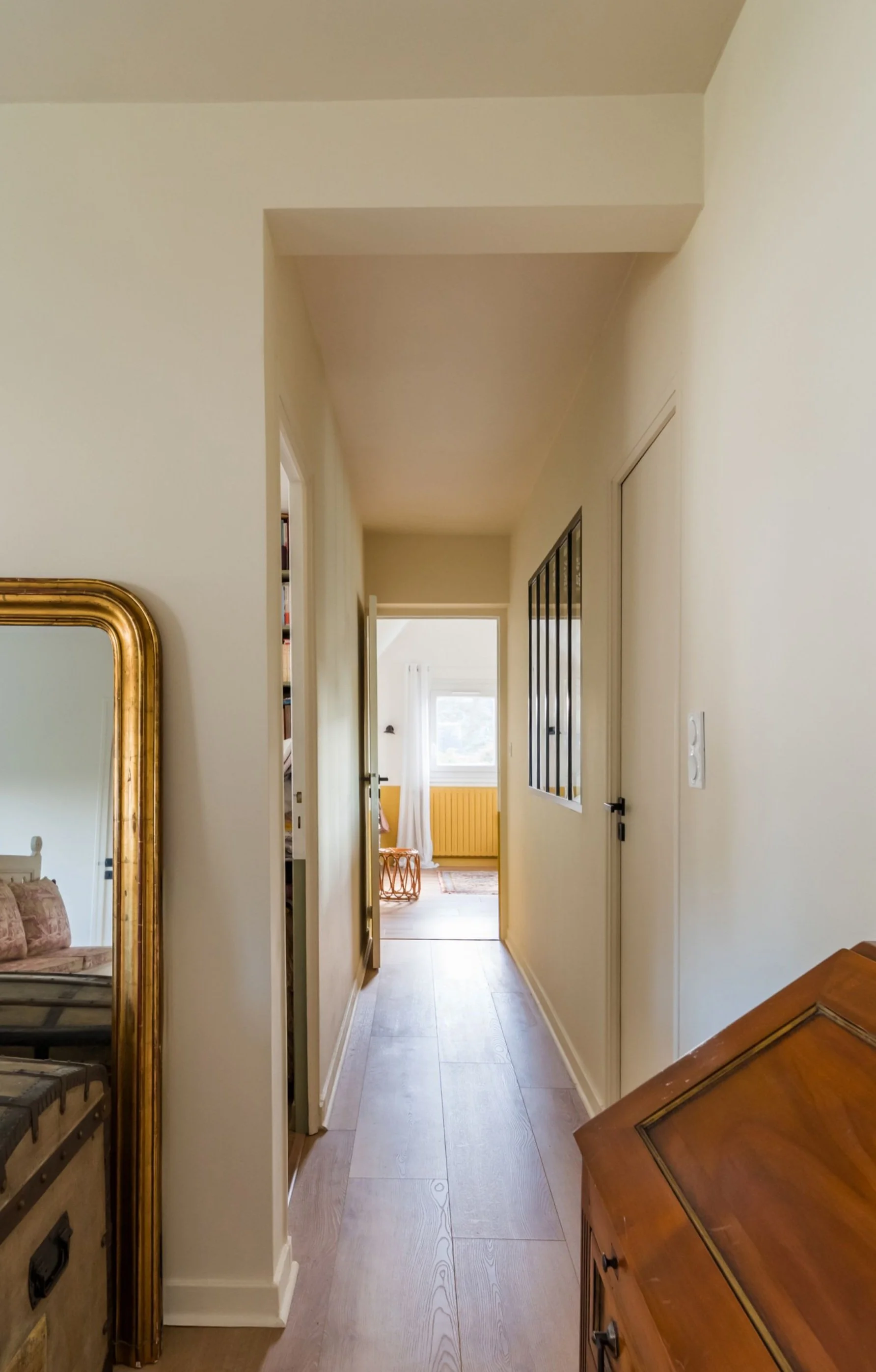 Vue d'un couloir dans une maison, avec un miroir doré sur la gauche, un mur à gauche avec un petit placard ou passage, et une pièce lumineuse à travers une porte ouverte en arrière-plan.