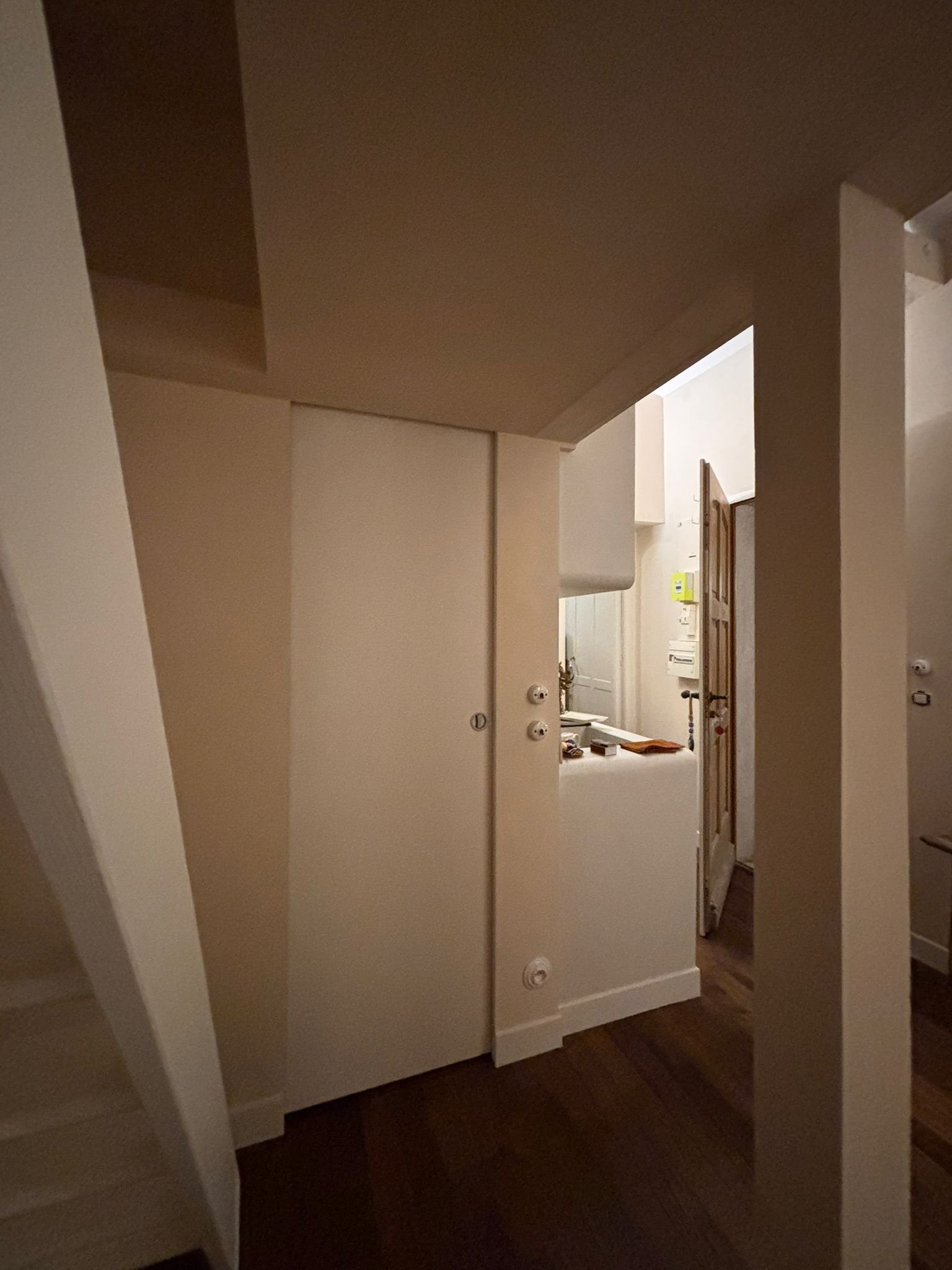 Une pièce intérieure avec des murs beiges, un plafond en bois foncé, une porte en bois à gauche, et un comptoir ou un meuble blanc. On peut voir une pièce à l'arrière avec un évier, et quelques objets sur le comptoir.
