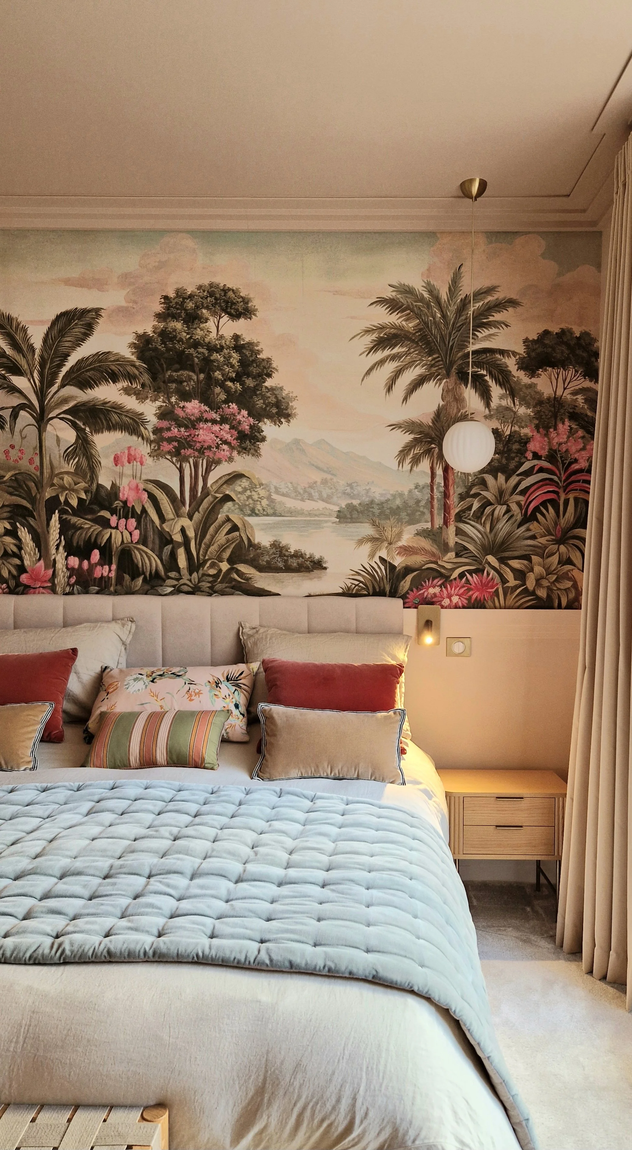 Chambre avec une tête de lit rembourrée, plusieurs coussins décoratifs aux motifs variés, une table de chevet en bois clair avec tiroirs, un mur peint avec un paysage de forêt tropicale et montagnes, un plafond avec un luminaire suspendu en forme de 