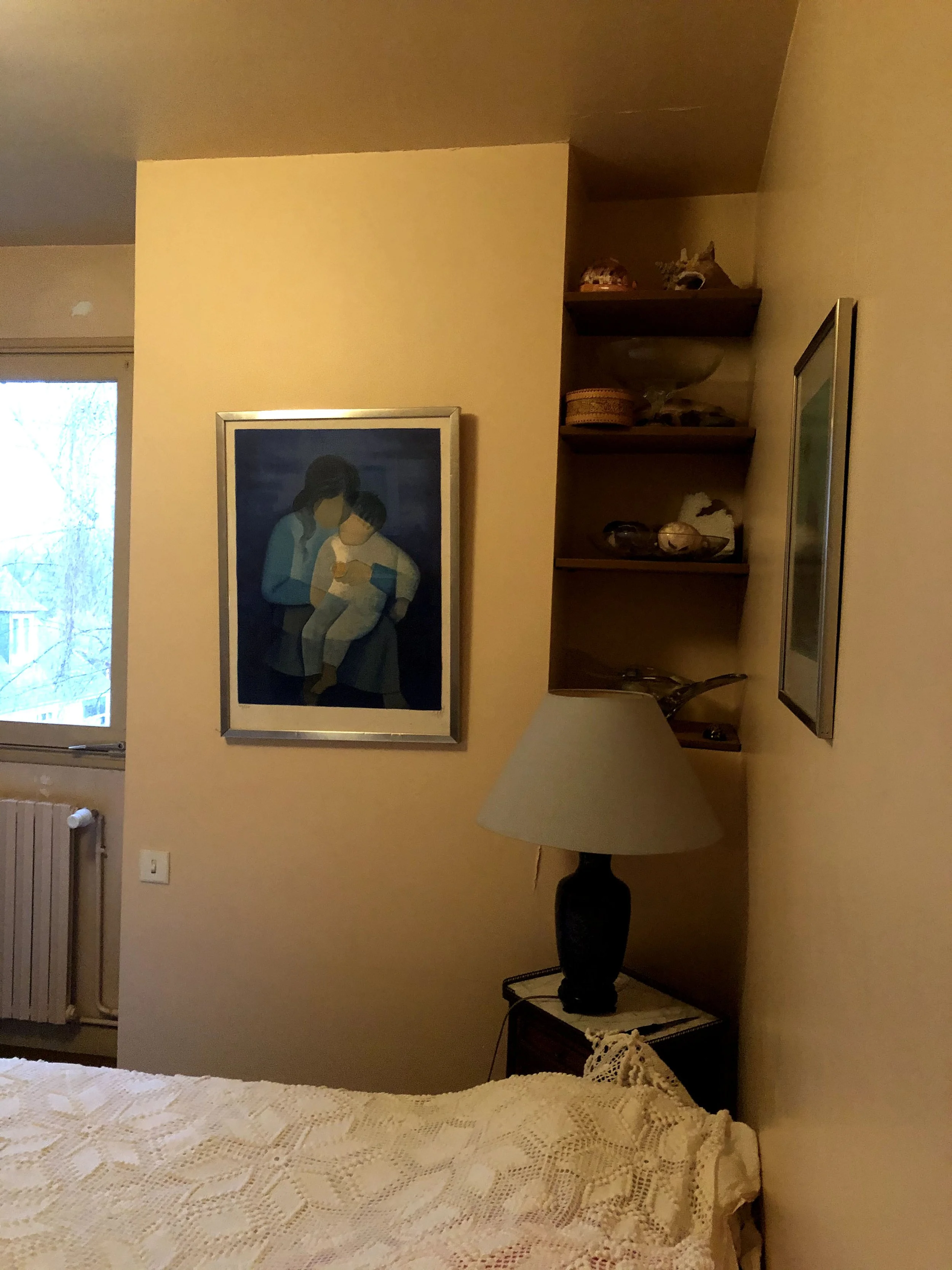 Intérieur d'une chambre avec un lit recouvert d'une couverture en crochet blanc, un poster d'une famille avec deux enfants, une étagère avec des objets décoratifs, une lampe sur une table, et une fenêtre avec vue sur des arbres.