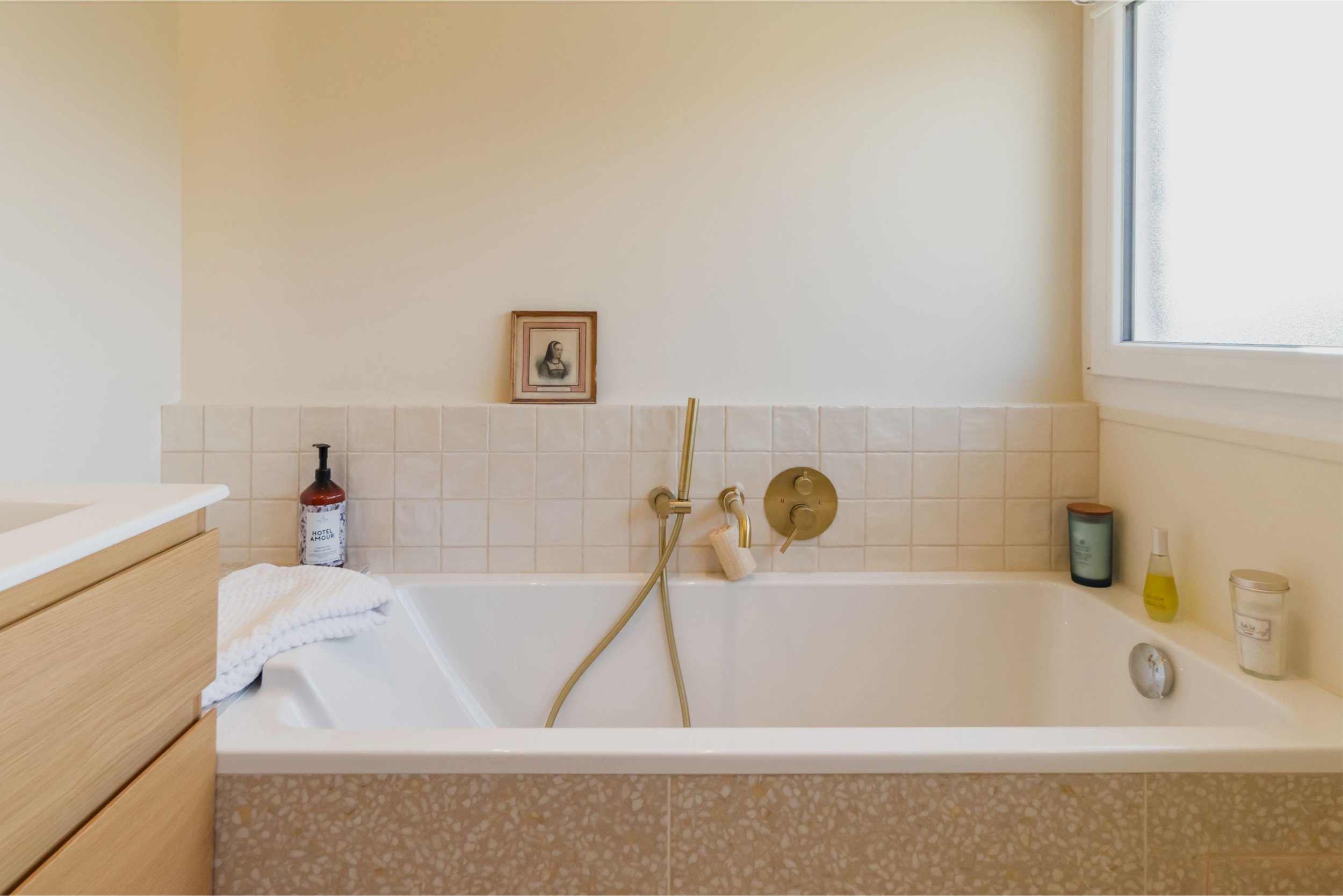 Une baignoire avec des produits de beauté et une serviette, dans une salle de bain claire avec un mur blanc, une fenêtre et un cadre avec une image d'une figure féminine.