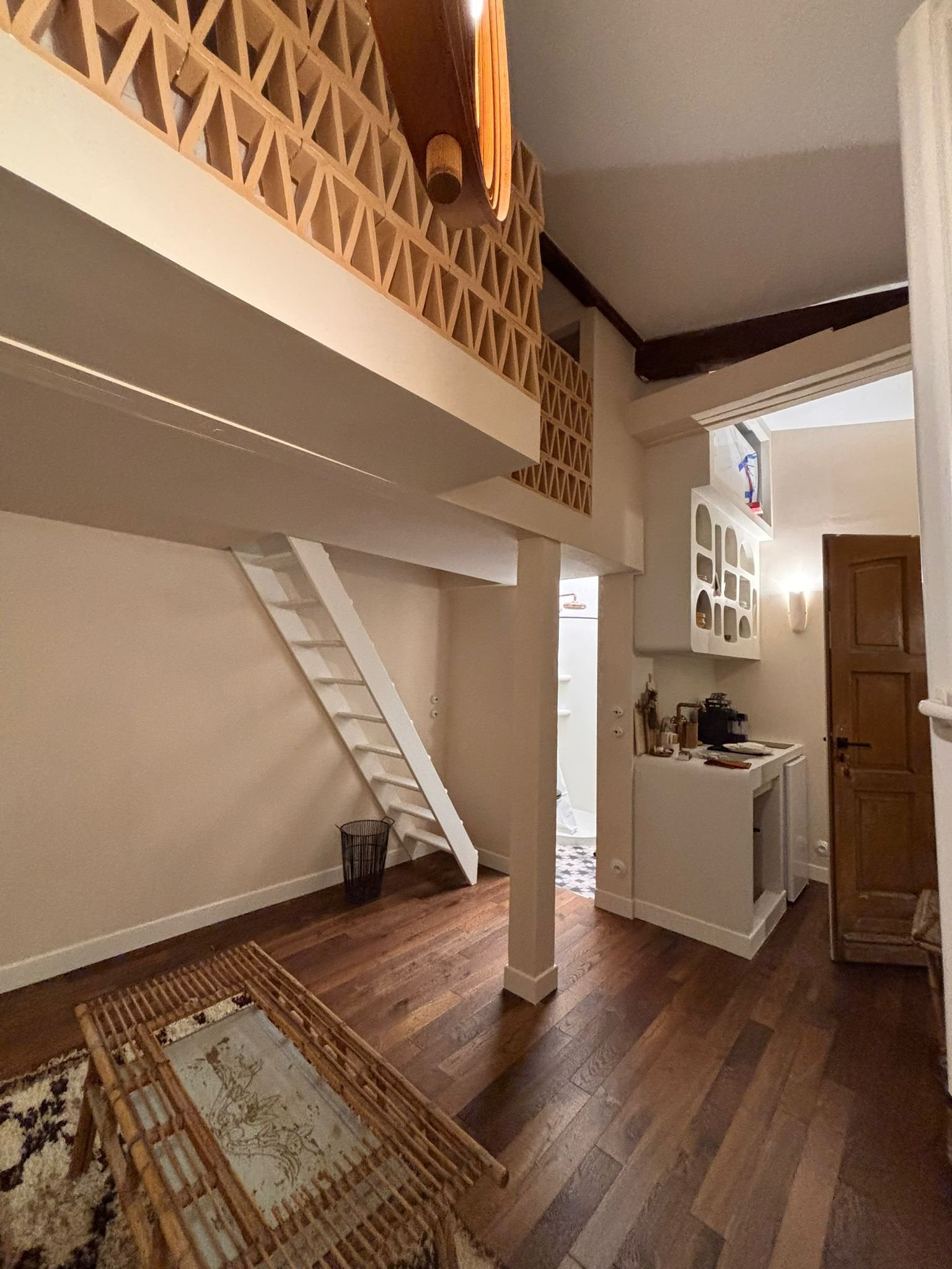 Intérieur d'une maison avec un plancher en bois, un escalier en bois éclairé par la lumière, un porte-manteau blanc, un meuble en bois avec des objets dessus, et un mur peint en beige.