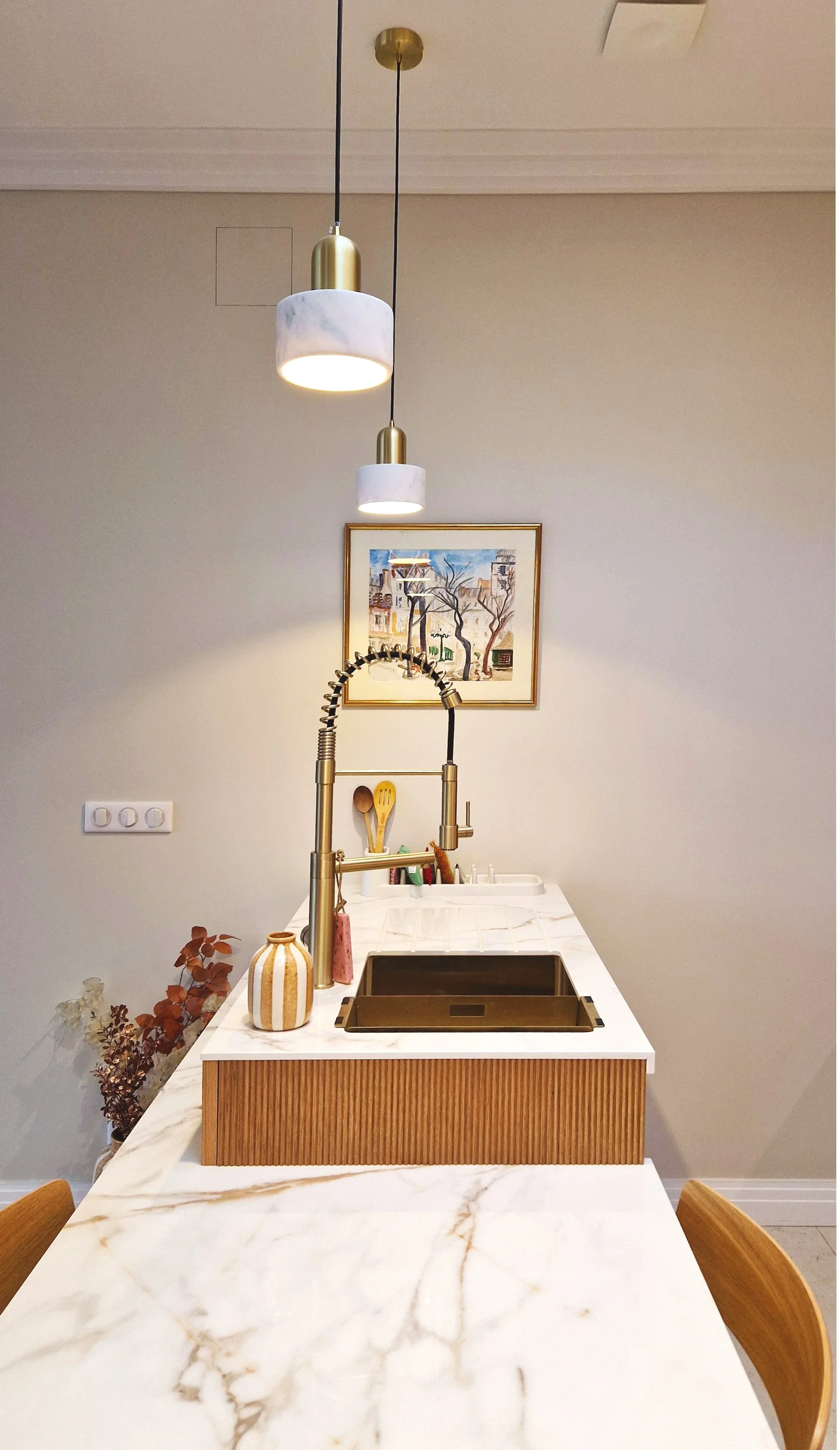 Cuisine moderne avec un plan de travail en marbre blanc, un évier doré, des décorations en pot à gauche, un tableau coloré sur le mur, et un plafond avec deux suspensions en marbre blanc et doré.