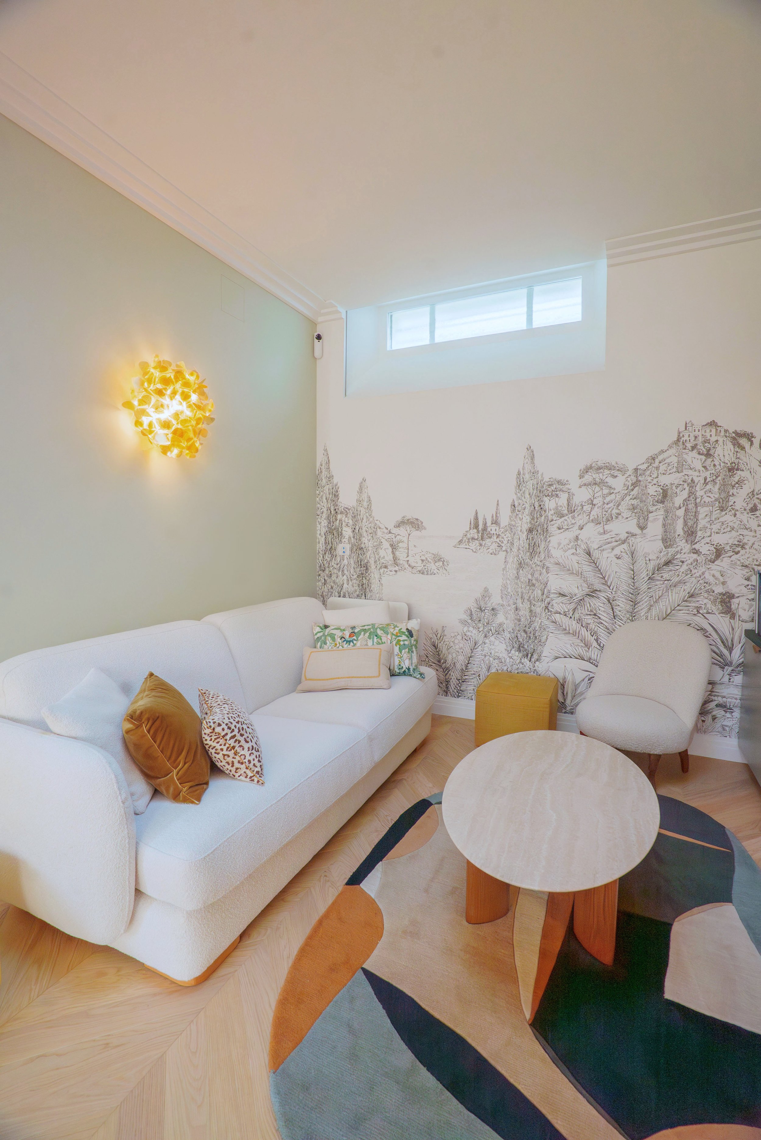 Salon moderne avec canapé blanc, coussins colorés, mur décoratif avec paysage, table basse, cheminée, fenêtre haute, tapis coloré, lampe murale