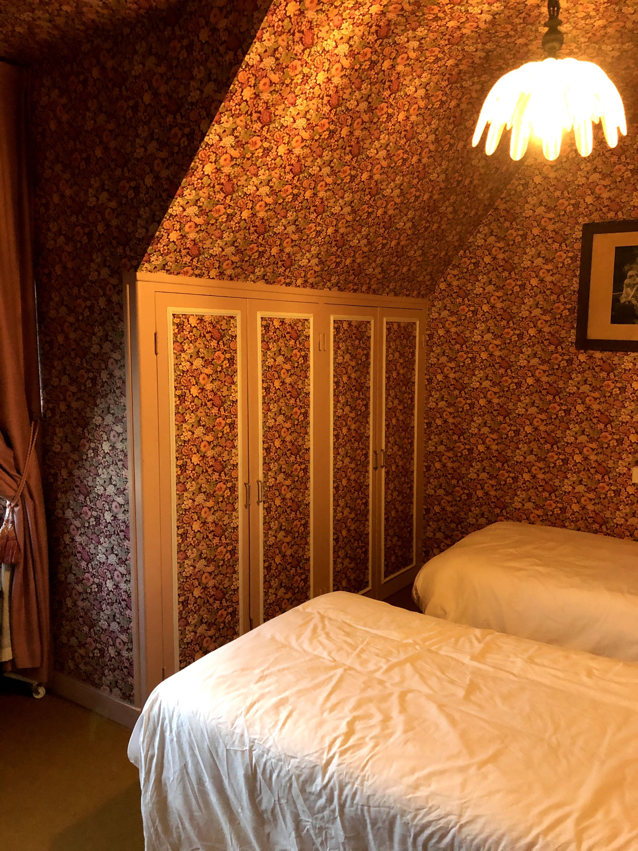 Chambre avec deux lits, murs et plafond recouverts de papier peint à motifs floraux orange et jaune, en particulier derrière l'armoire et le lit.