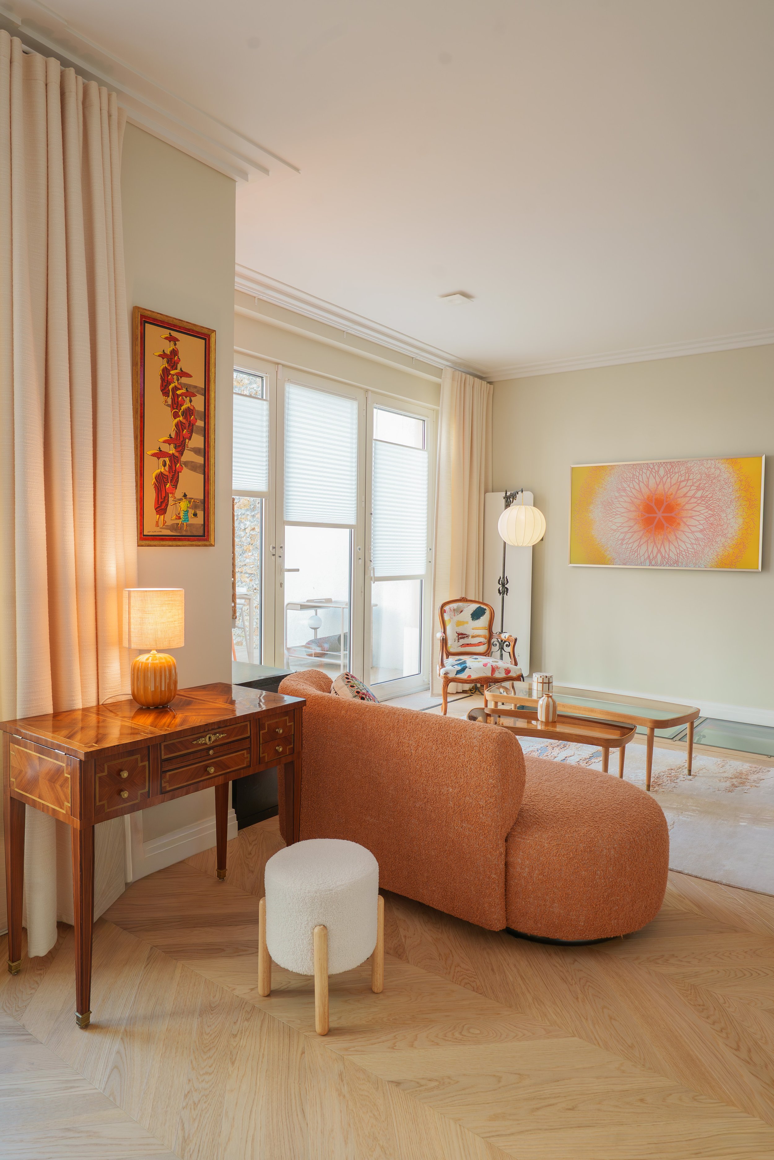 Salon avec un canapé orange, une table basse en bois, une lampe de table orange, un fauteuil en bois avec un coussin coloré, des œuvres d'art sur les murs, et de grandes fenêtres avec rideaux clairs. 