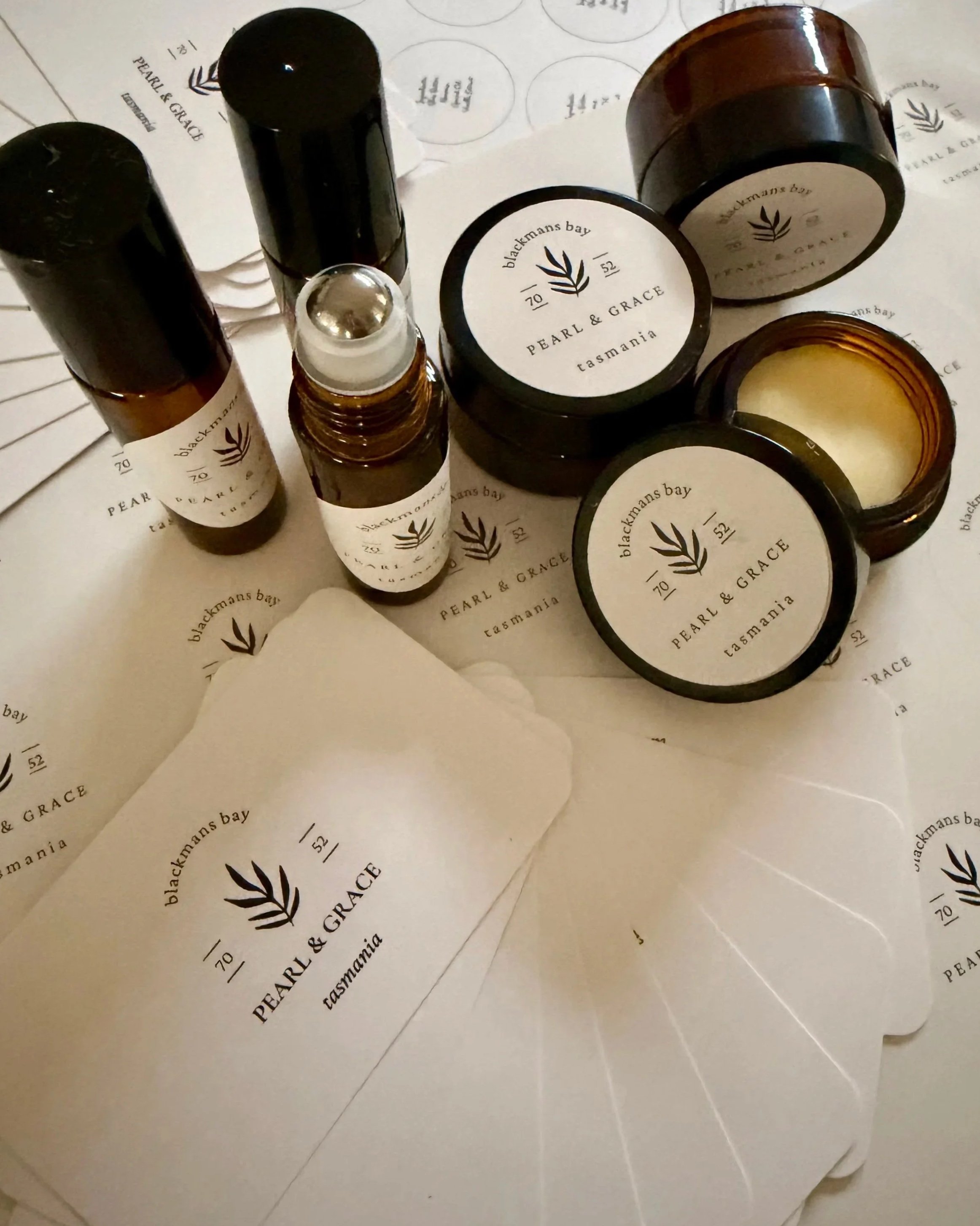 A collection of skincare or beauty product amber glass roller perfume bottle lip balm pots with white labels black writing that reads ' pearl & grace blackmails bay tasmania,' placed on printed paper and tissue paper with matching branding.