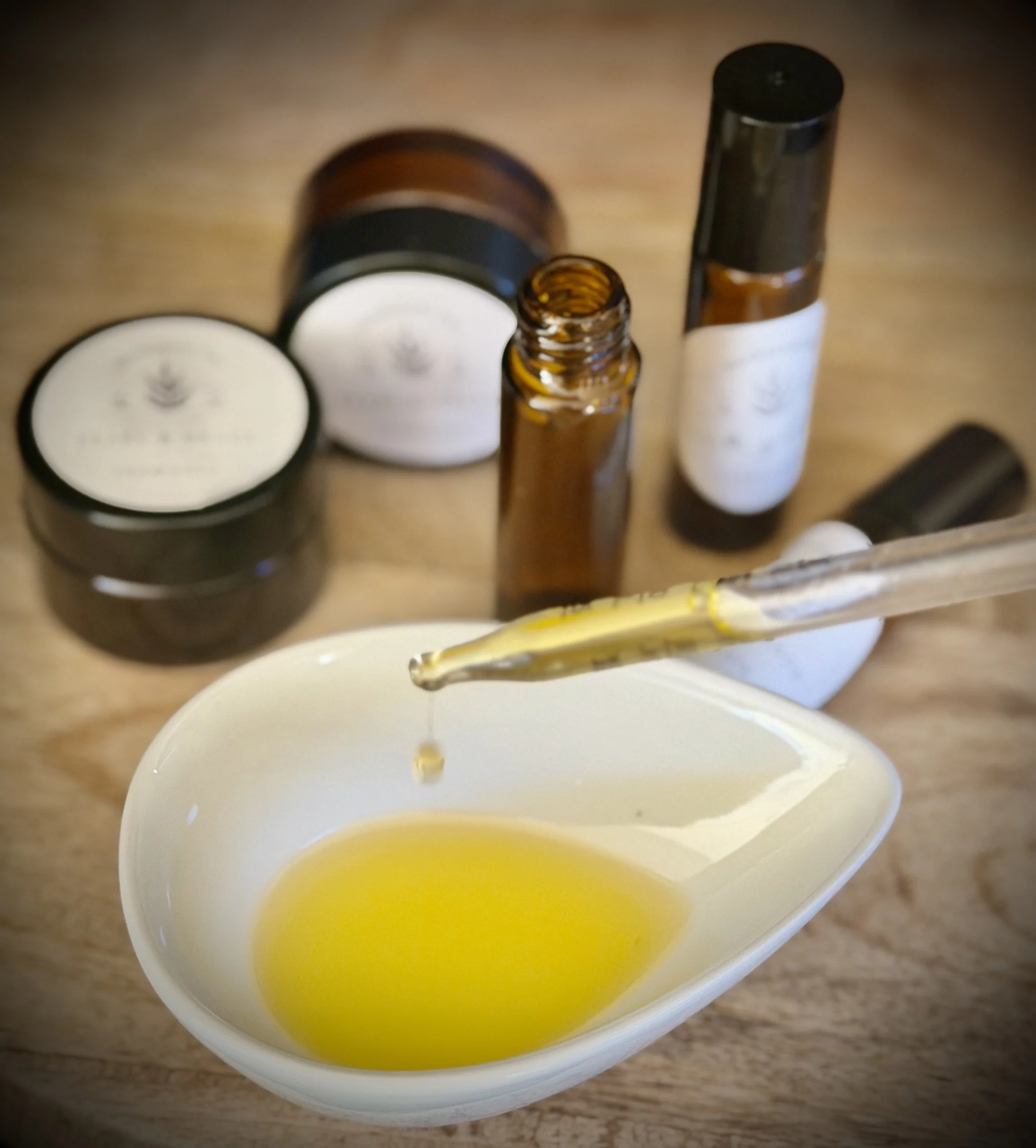 Close-up of a small white dish with yellow oil, with several bottles and jars in the background, possibly containing essential oils or skincare products.