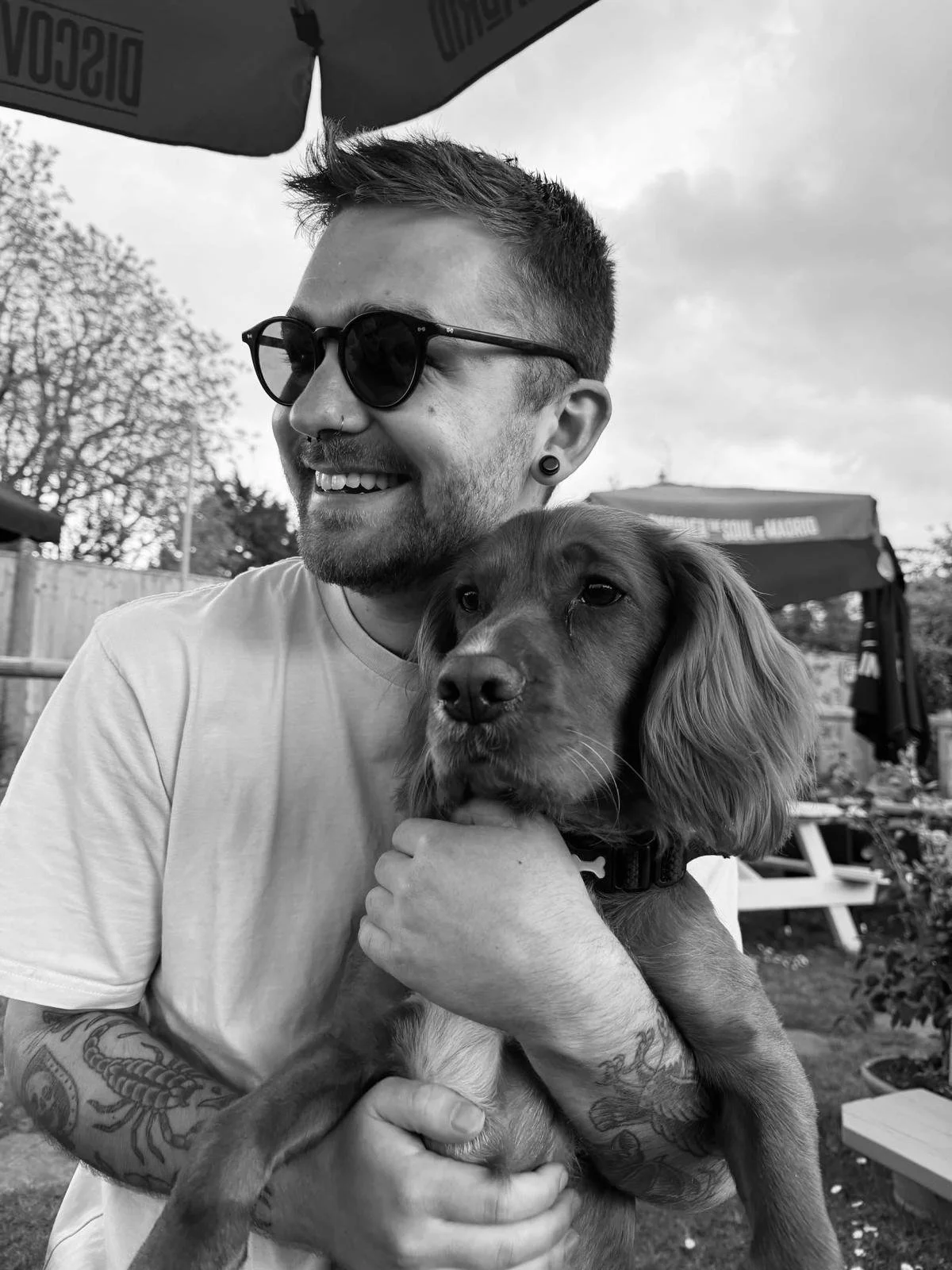 A smiling young man with sunglasses and tattoos on his arm holding a medium-sized dog with floppy ears outdoors.
