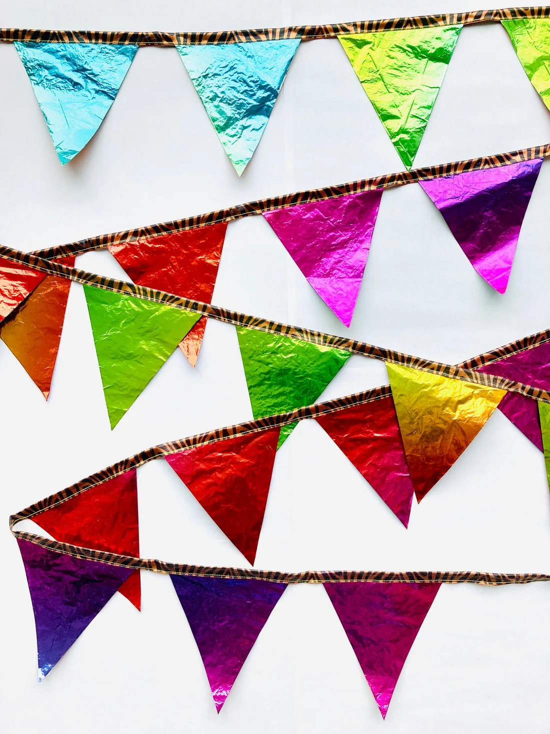 Multicoloured party bunting banners made of shiny recycled balloons, hanging on striped ribbons against a white background.